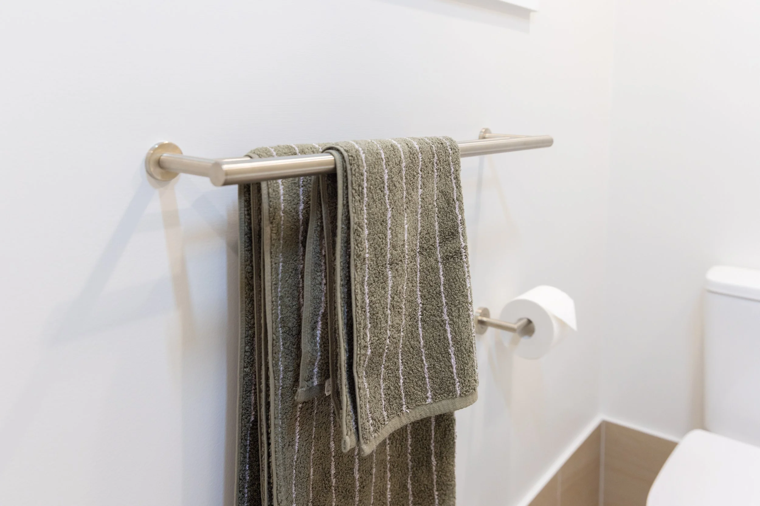 Bathroom wall with a metal towel rack holding two hanging gray towels with white stripes, and a toilet paper roll on a metal holder below.