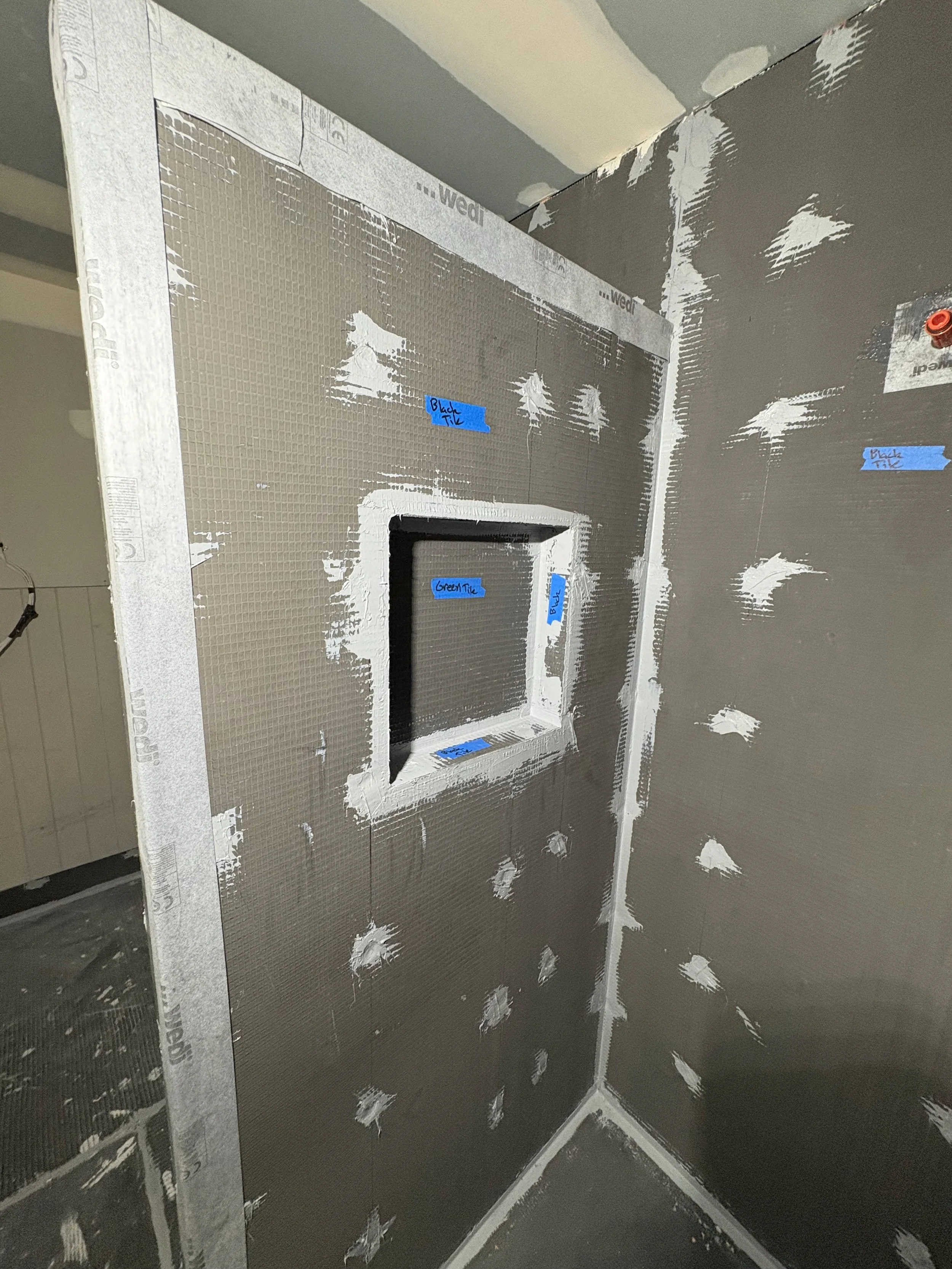 Drywall installation in progress on a corner of a room, with a small square window cutout. The drywall has labels for different tile colors: blue labels for black and green tiles.