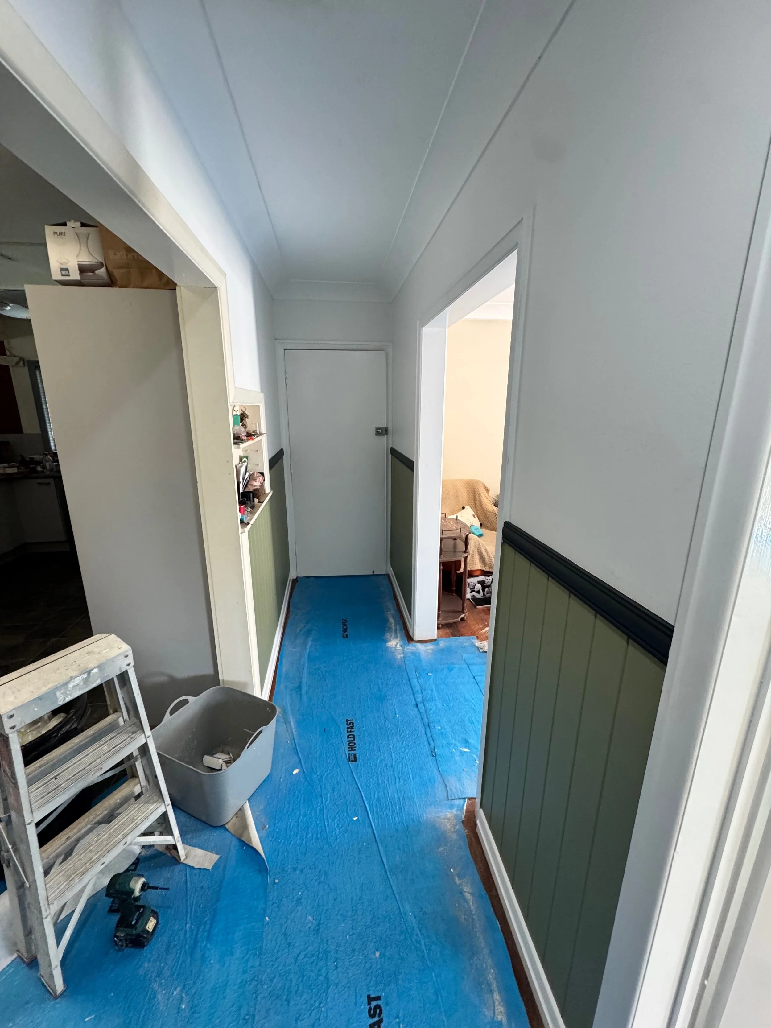 A hallway under renovation with blue protective floor covering, a small ladder, a plastic bin, and construction tools, leading to a closed door and a living room area with a sofa.