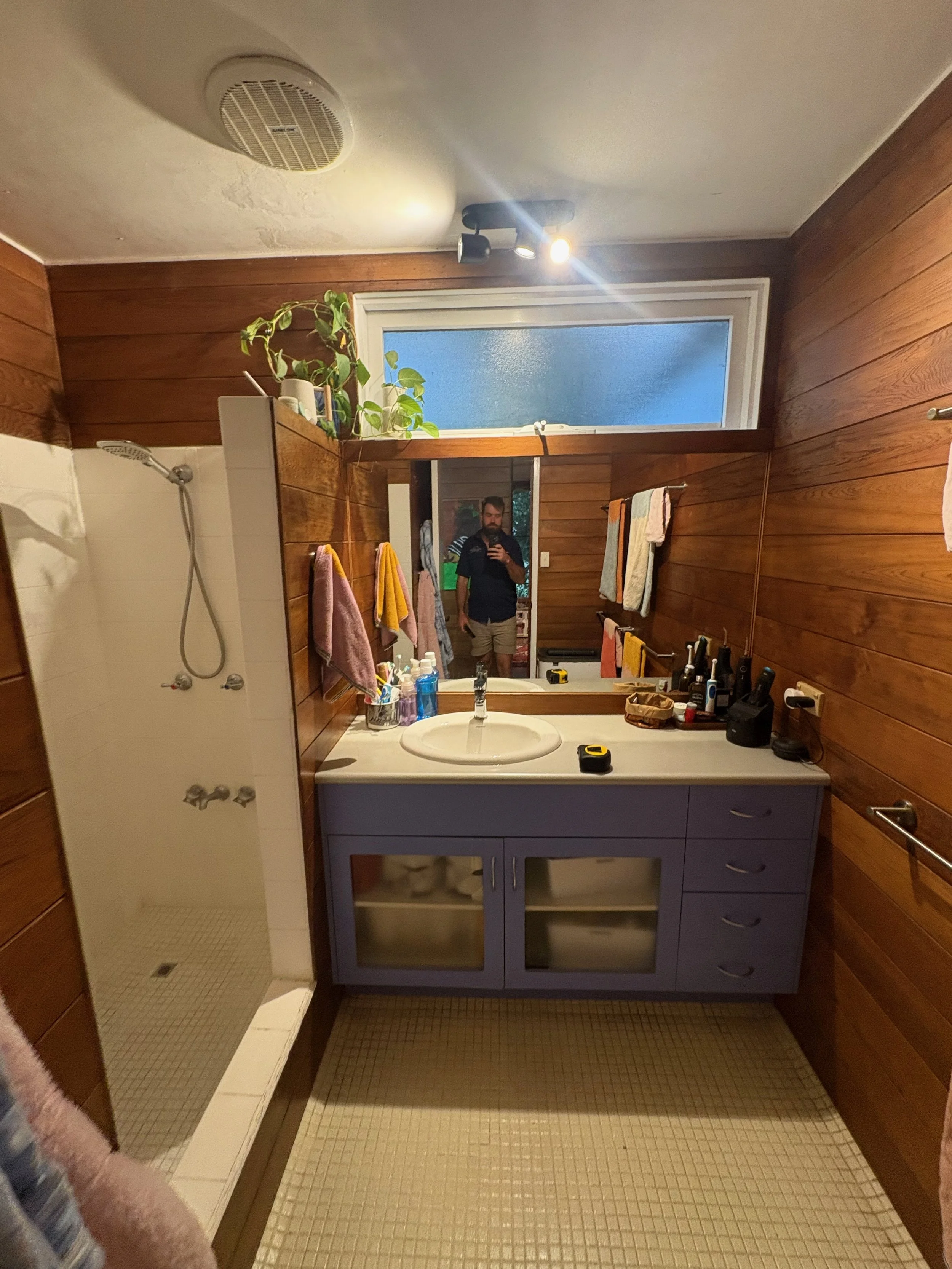 A small bathroom with wooden-paneled walls, a shower stall on the left, a purple vanity with a white sink in the center, a large mirror above the vanity, and a frosted window above the mirror. Various toiletries and items are on the vanity, with towe