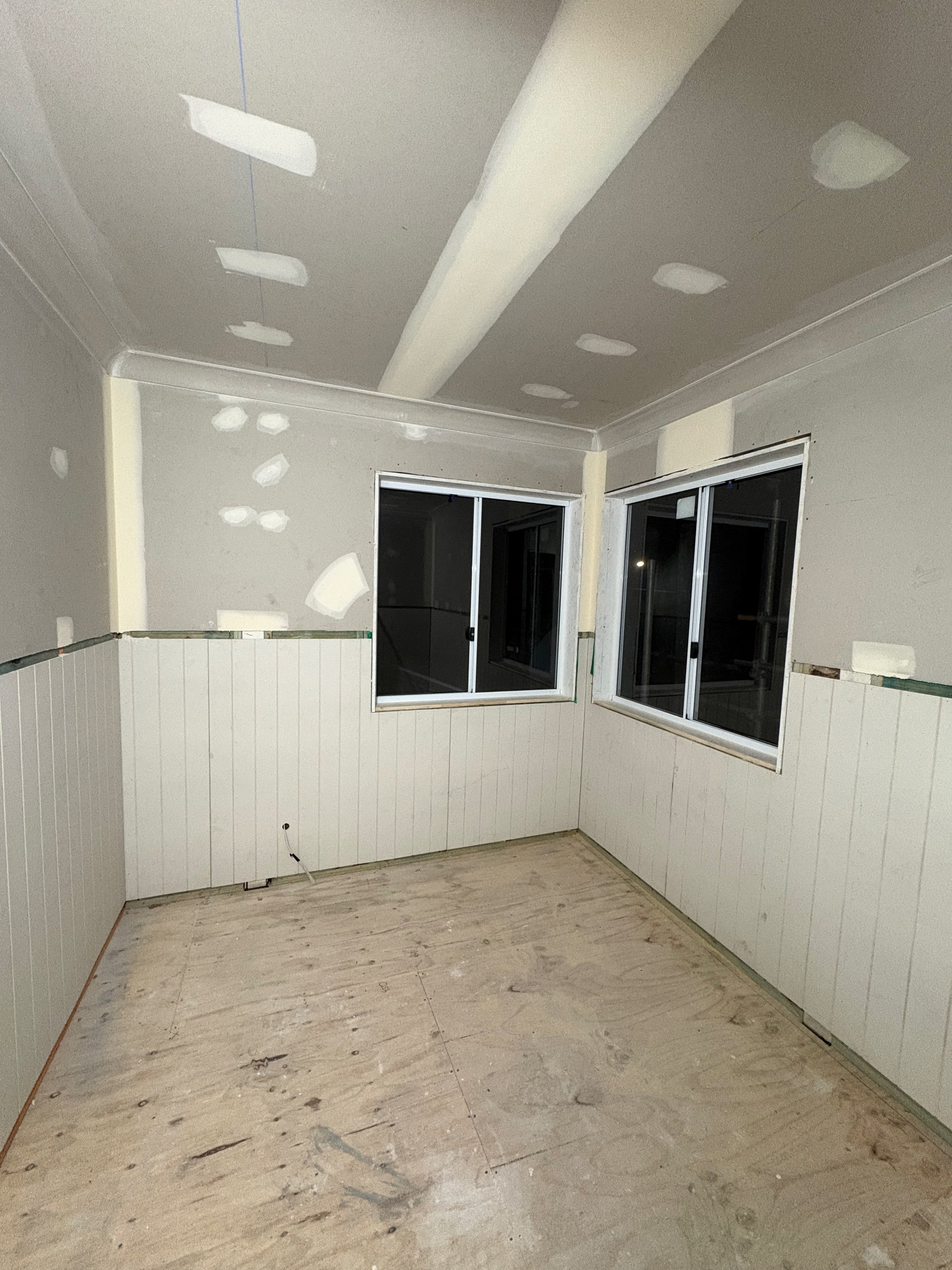 Unfinished room with drywall and beadboard paneling, two windows, and plywood flooring, likely under renovation.