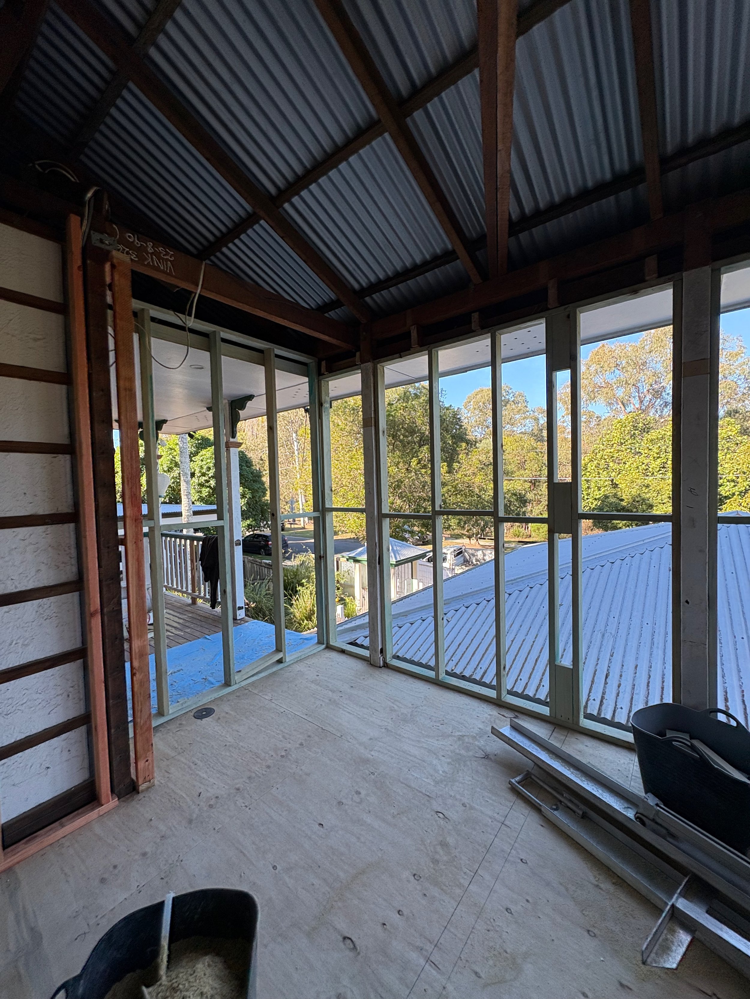 Construction of a screened-in porch with view of trees and rooftops, showing framing, partial flooring, and a black bucket in the foreground.