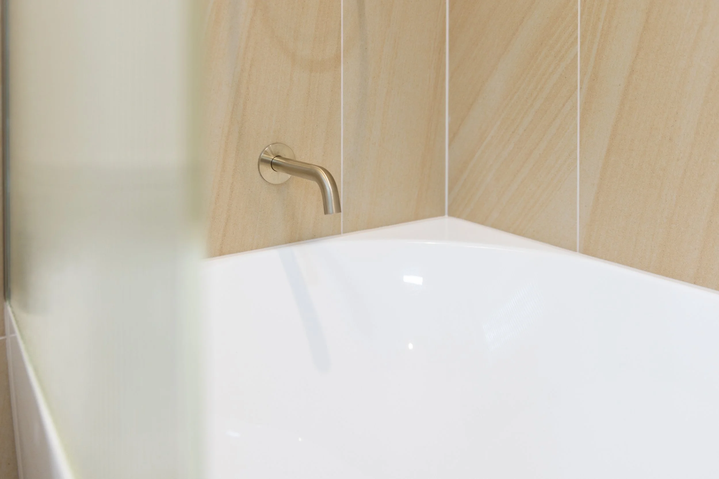 Close-up of a white bathtub with a wall-mounted metallic faucet in a bathroom with beige tiled walls.