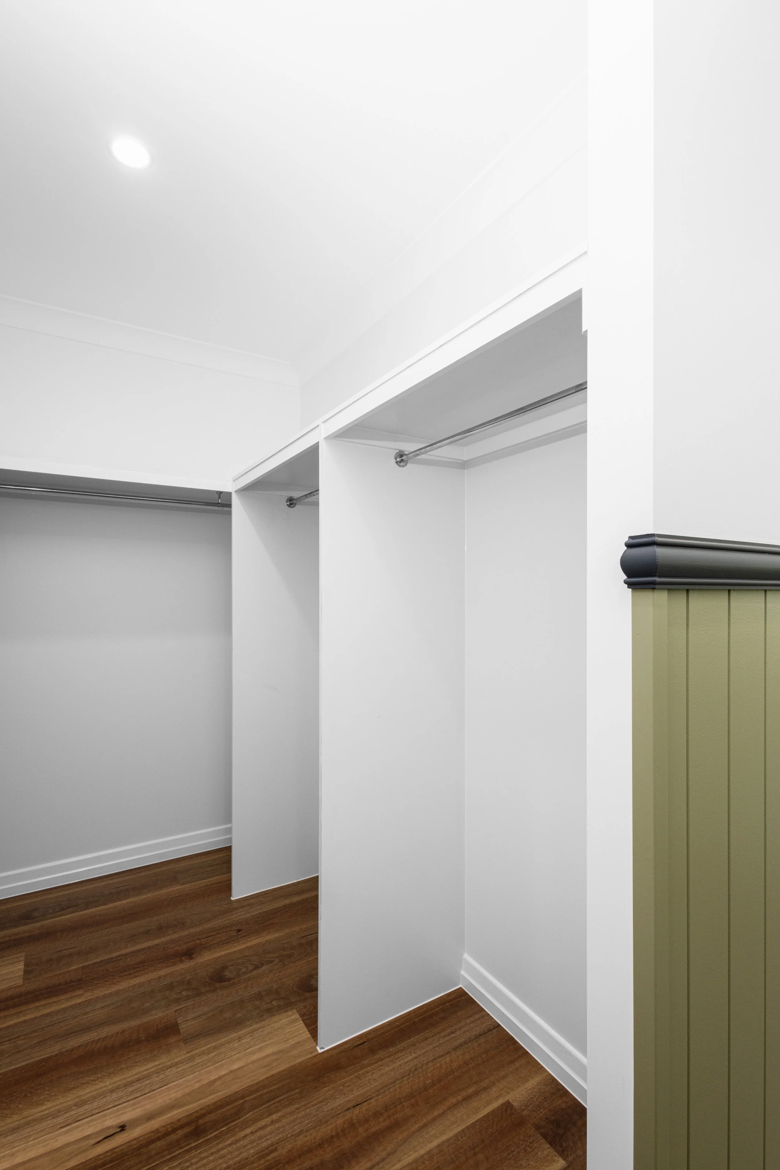 Empty white closet with open space for hanging clothes, wood flooring, and a small section of green wall paneling.
