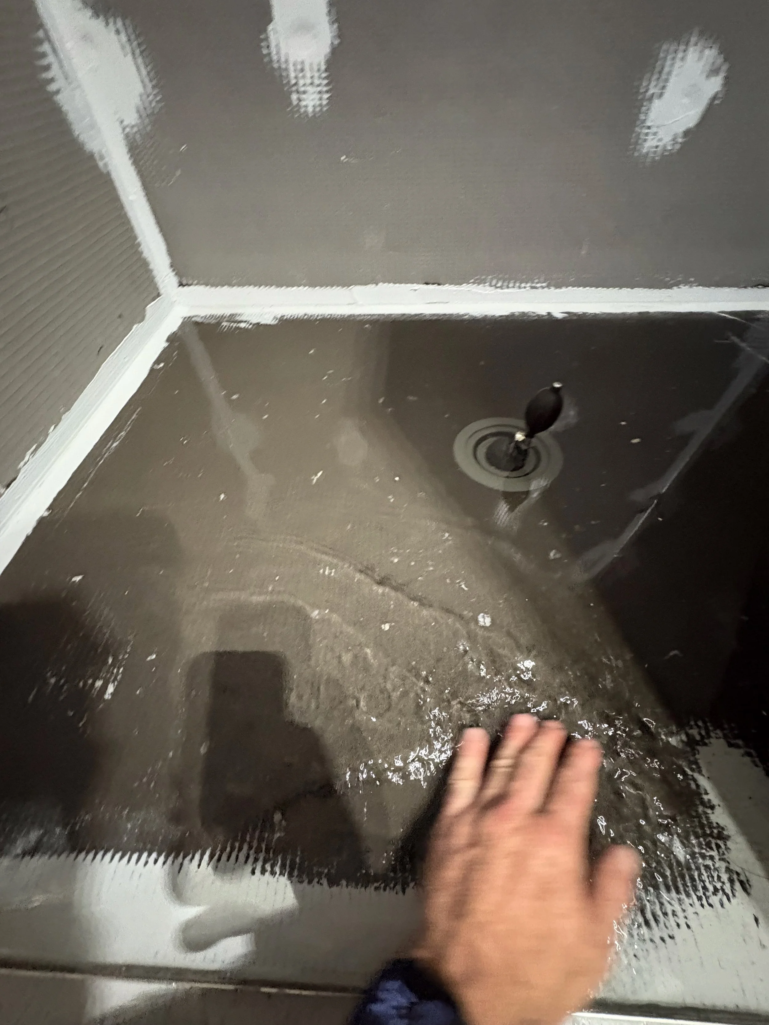 A person is working on installing or repairing a shower or bathtub, with a hand touching the surface and plumbing fixtures visible in the corner.