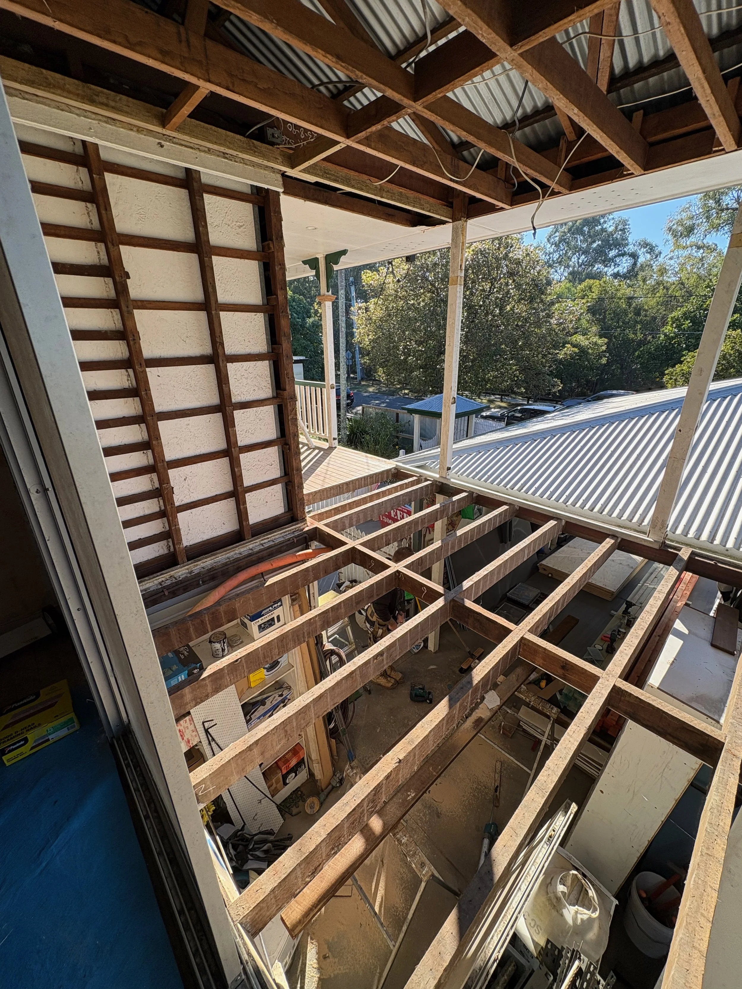 Construction site of a building with a wooden framework, open to the outside, showing trees and rooftops.