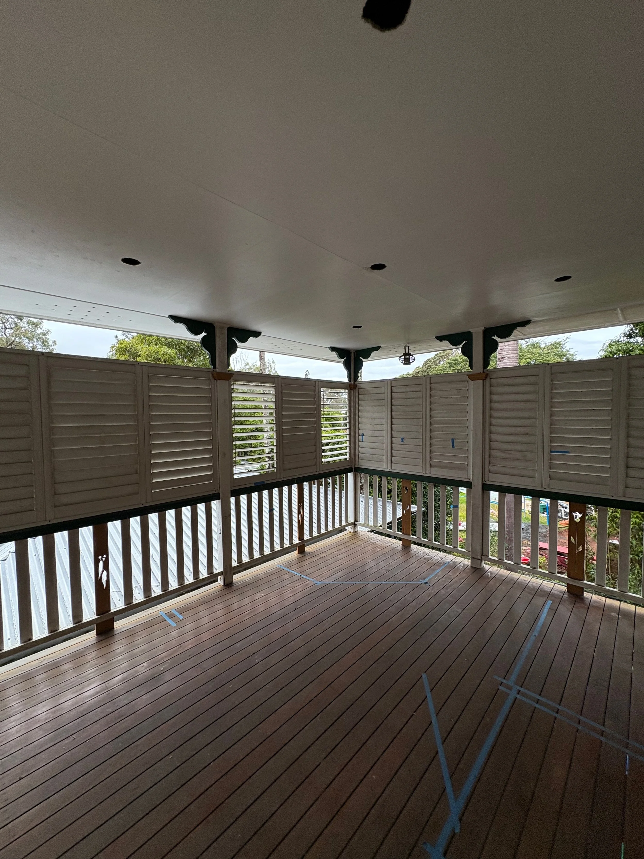 Undeveloped balcony with wooden flooring, partly covered ceiling, and white railings and shutters, with some marked tape on the floor for construction or decoration, and trees visible outside.
