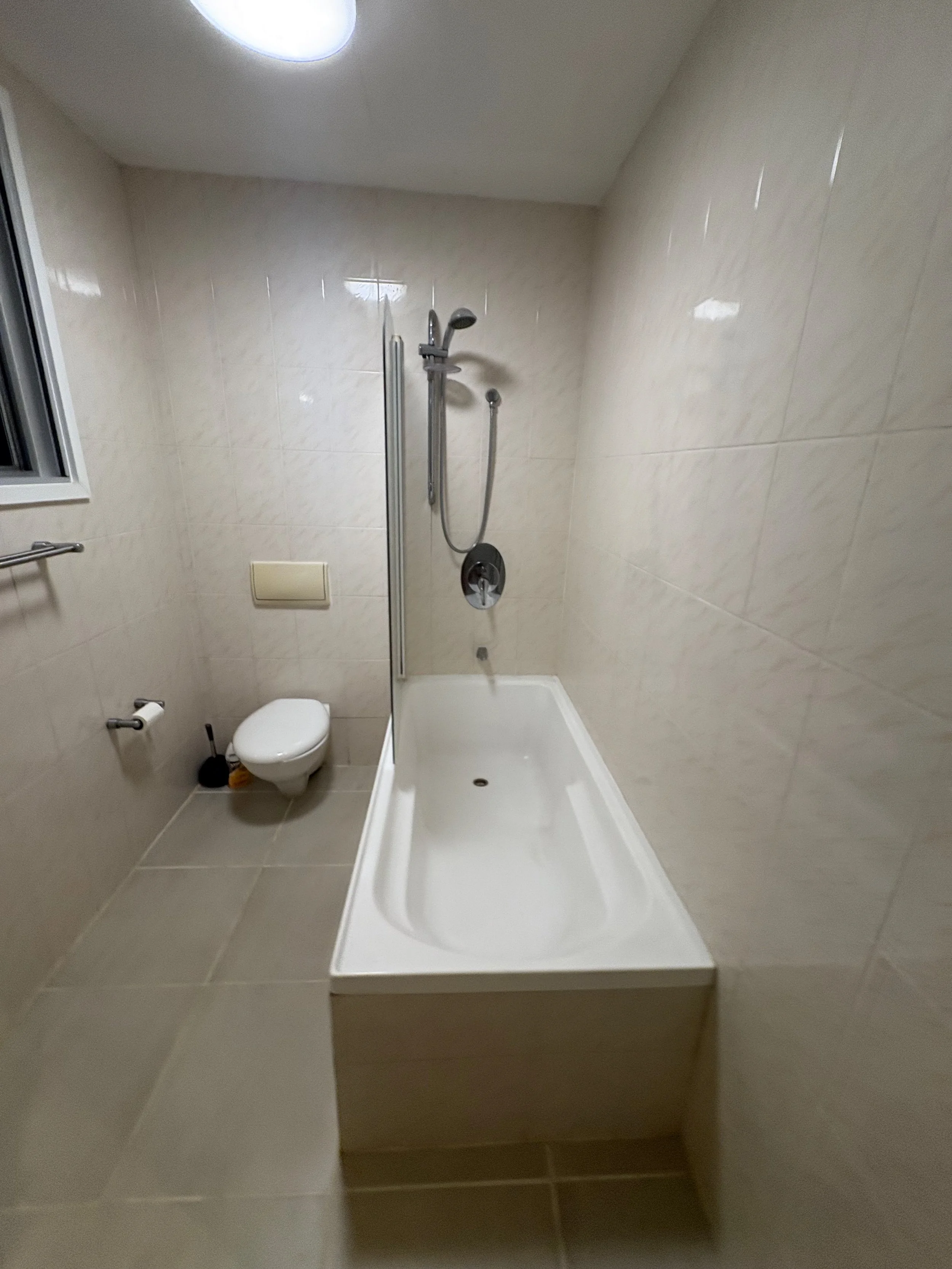 A small bathroom with beige tiled walls and floor, featuring a shower, a toilet, a window, and a toilet paper holder.