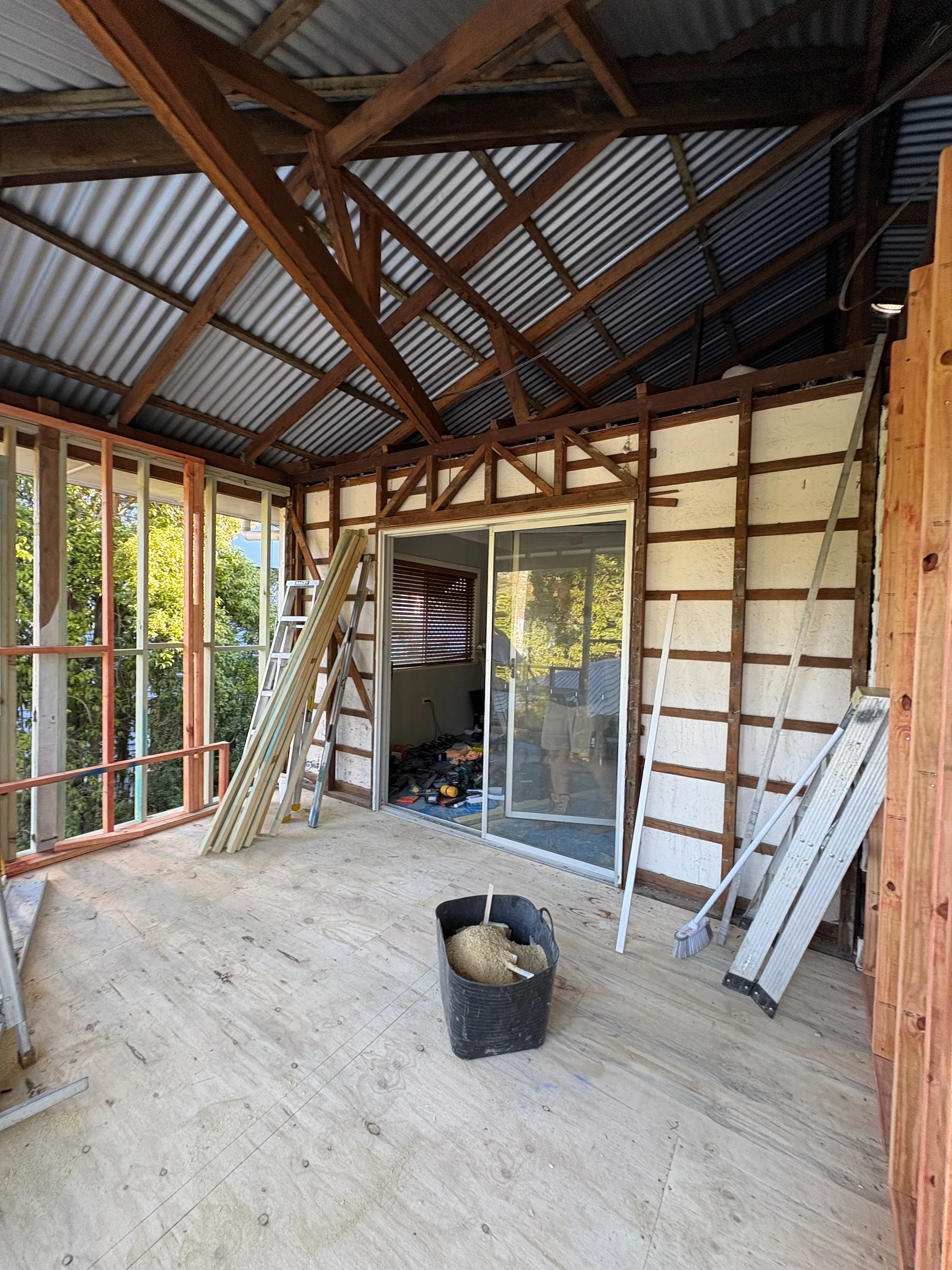Construction site of a room with exposed wooden framing and sliding glass door, construction tools, ladder, and construction materials on the unfinished floor.