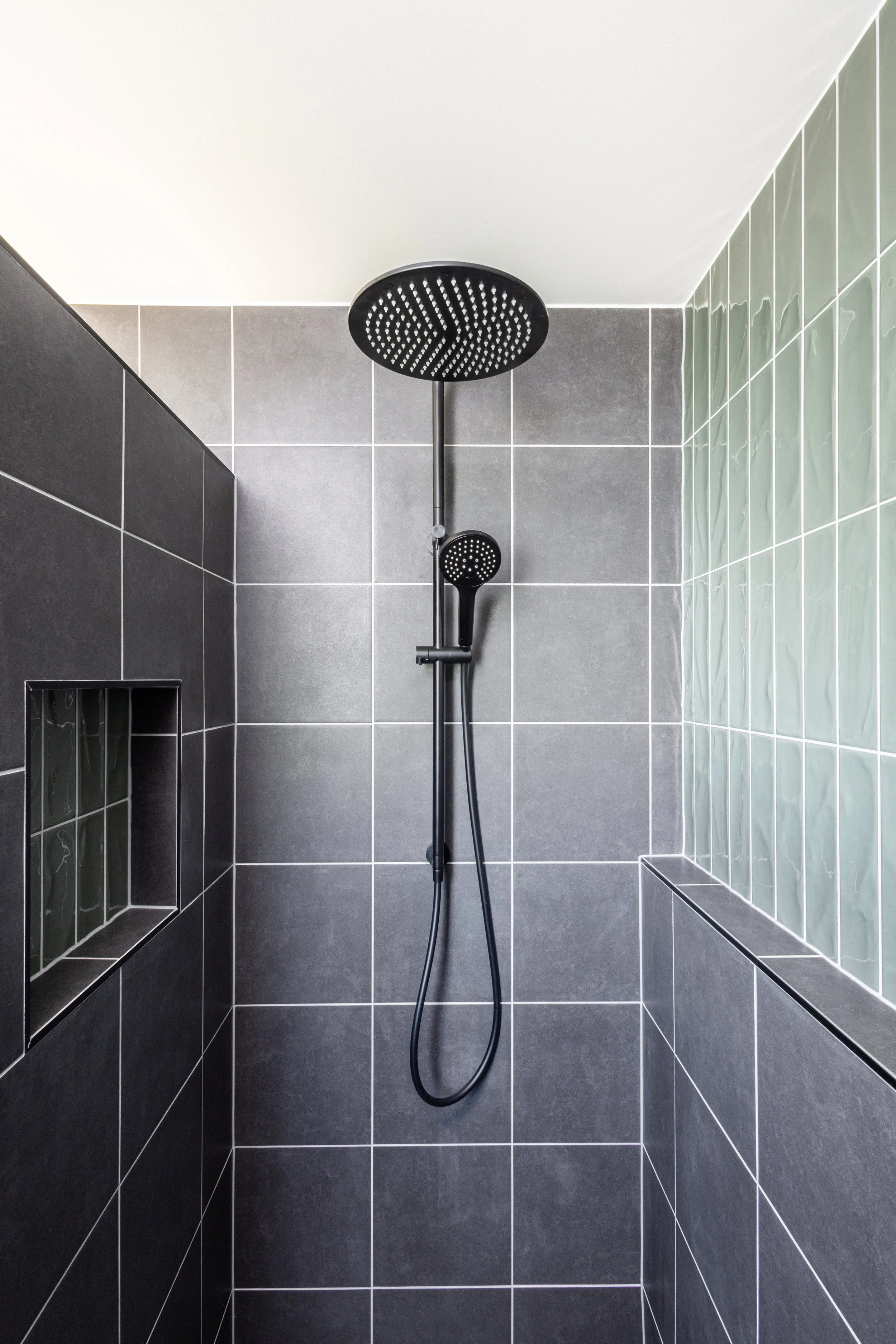 Modern shower with black showerhead and handheld sprayer on dark gray tiled walls, with built-in niche and green-tiled accent wall.