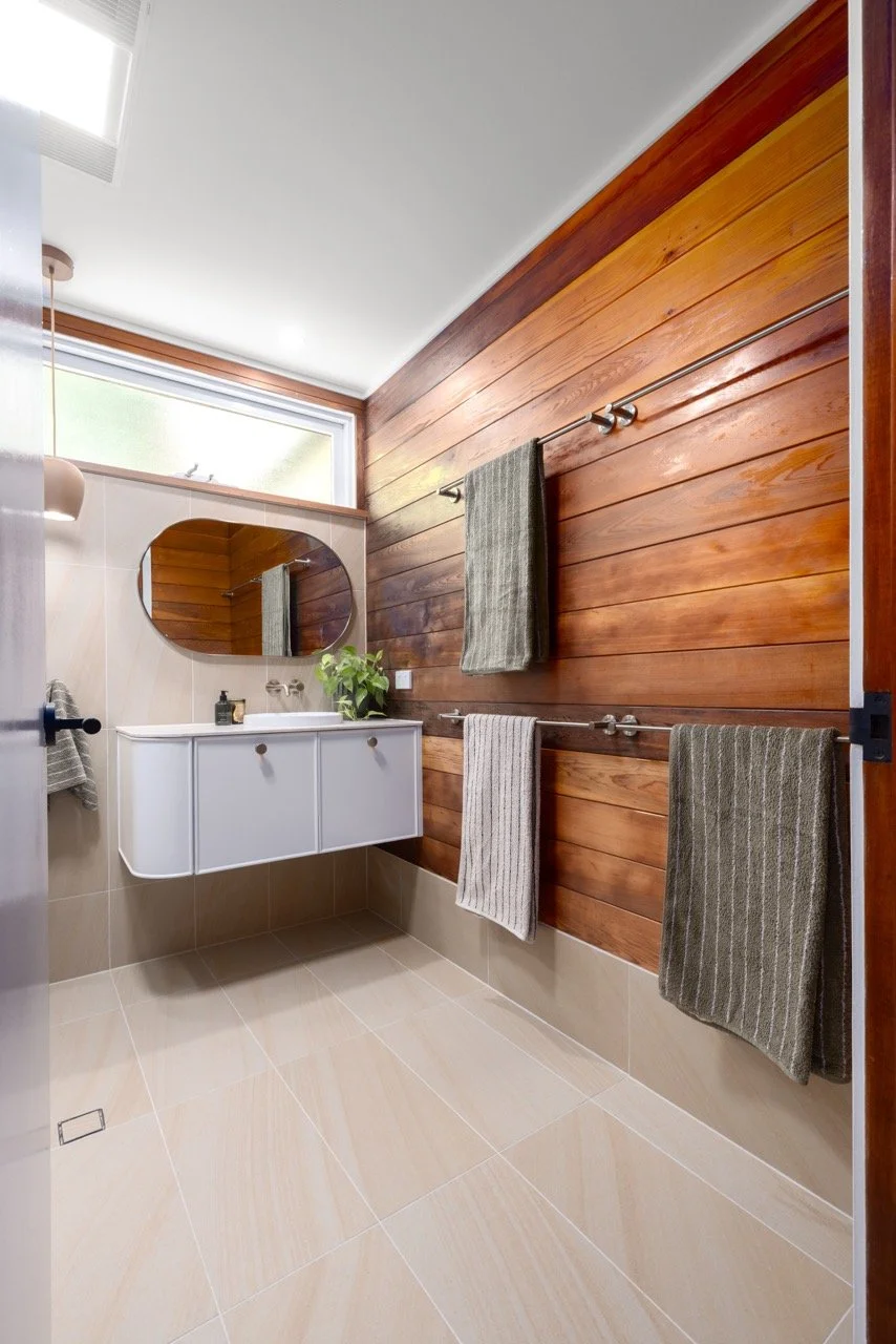 Modern bathroom with wood-paneled wall, beige tile floor, white wall-mounted vanity with round mirror, green plant, and three hanging towels.