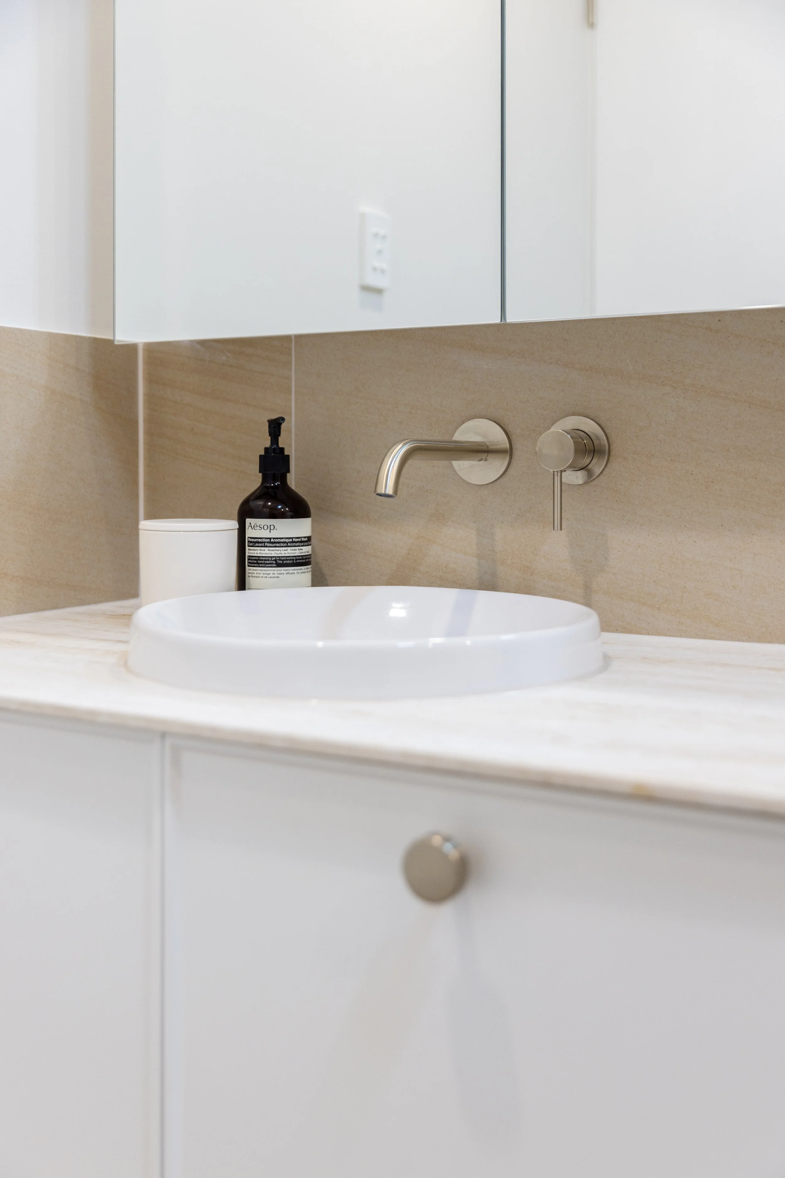 Bathroom sink with a white basin, soap dispenser, and small container on a beige countertop, with a mirror above and a white cabinet below.