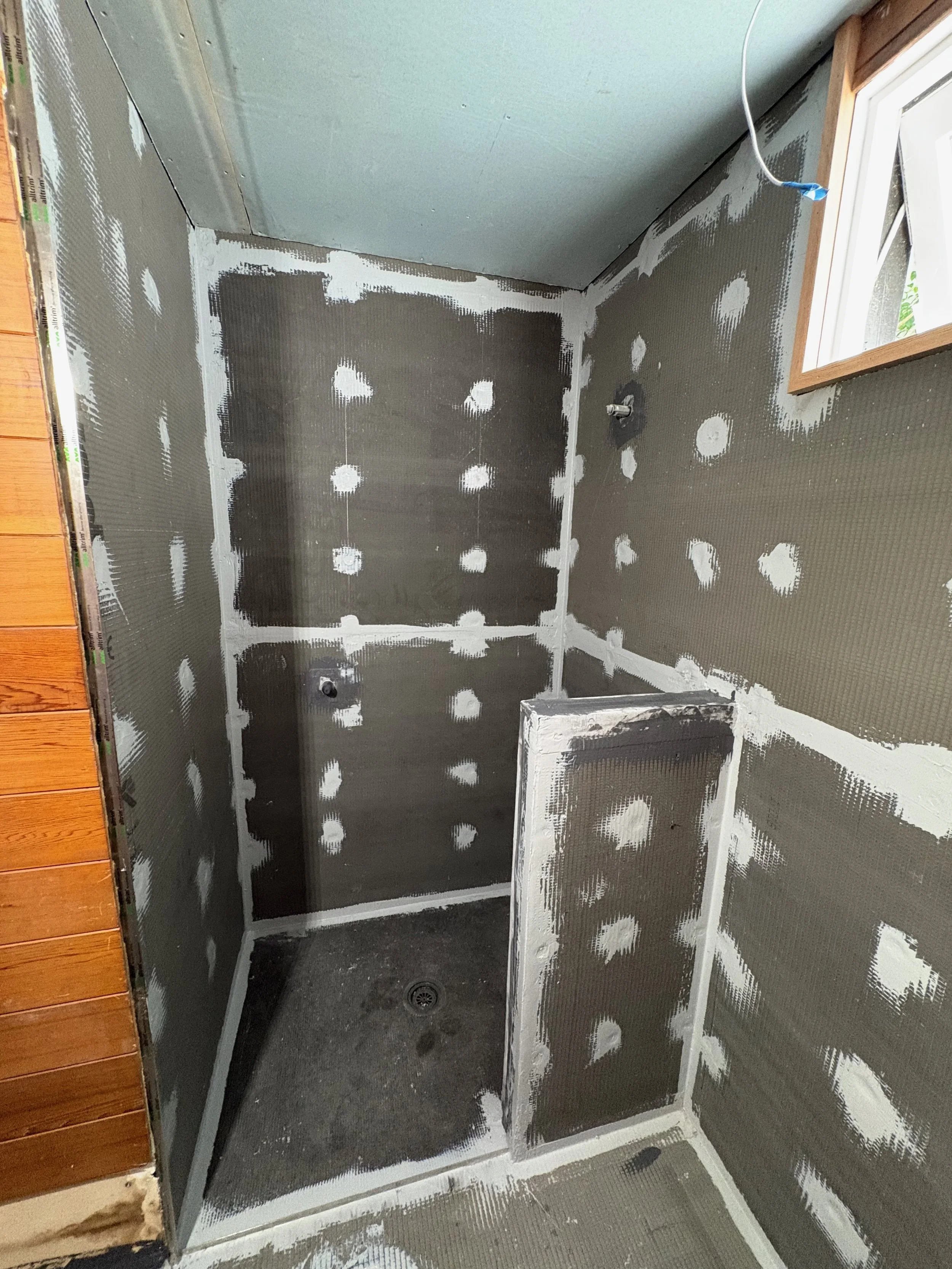 Shower stall under construction with unfinished drywall, visible patching, and wiring, with a small window and drain on the floor.
