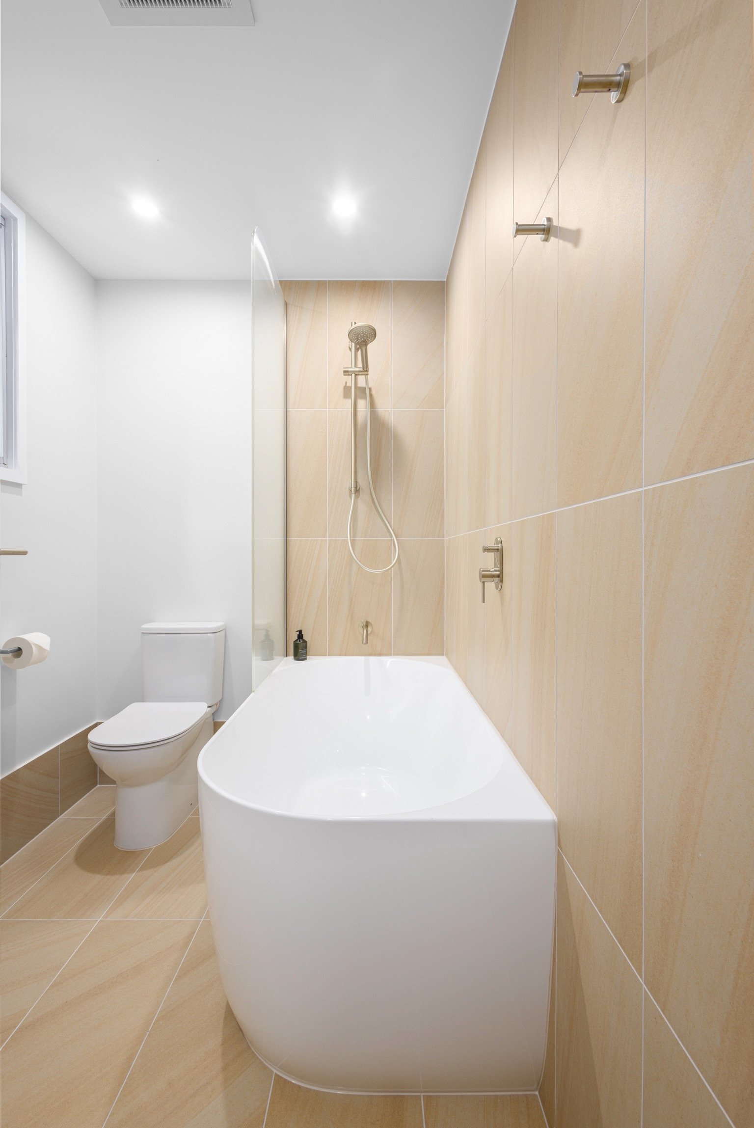 A minimalist bathroom with a white bathtub, a toilet, and a beige tiled wall with a showerhead and hooks.