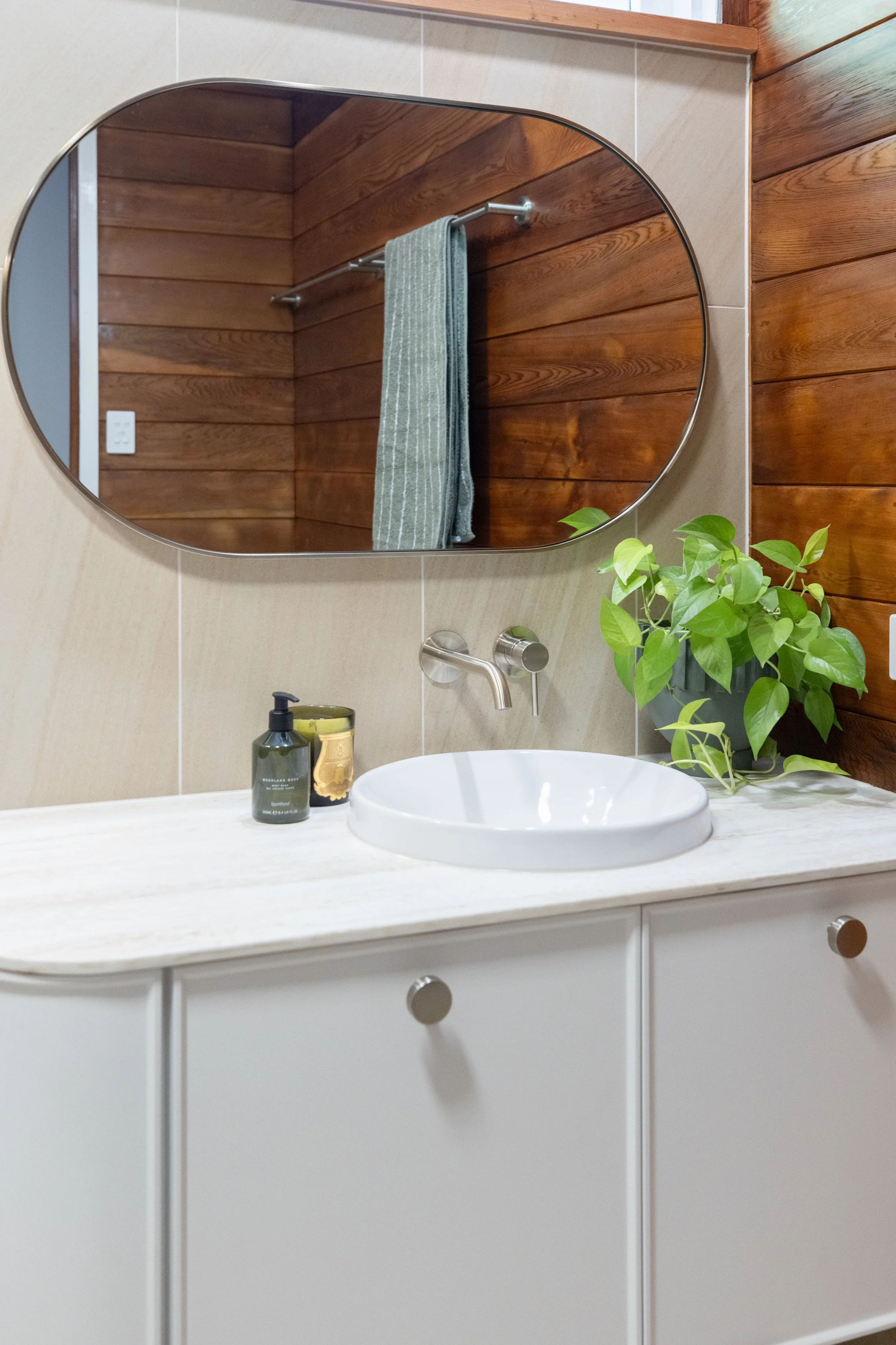 A bathroom sink with a white basin on a light-colored vanity, a soap dispenser, a candle, a plant, a water faucet, a mirror, a towel, wooden paneled walls, and a light switch.