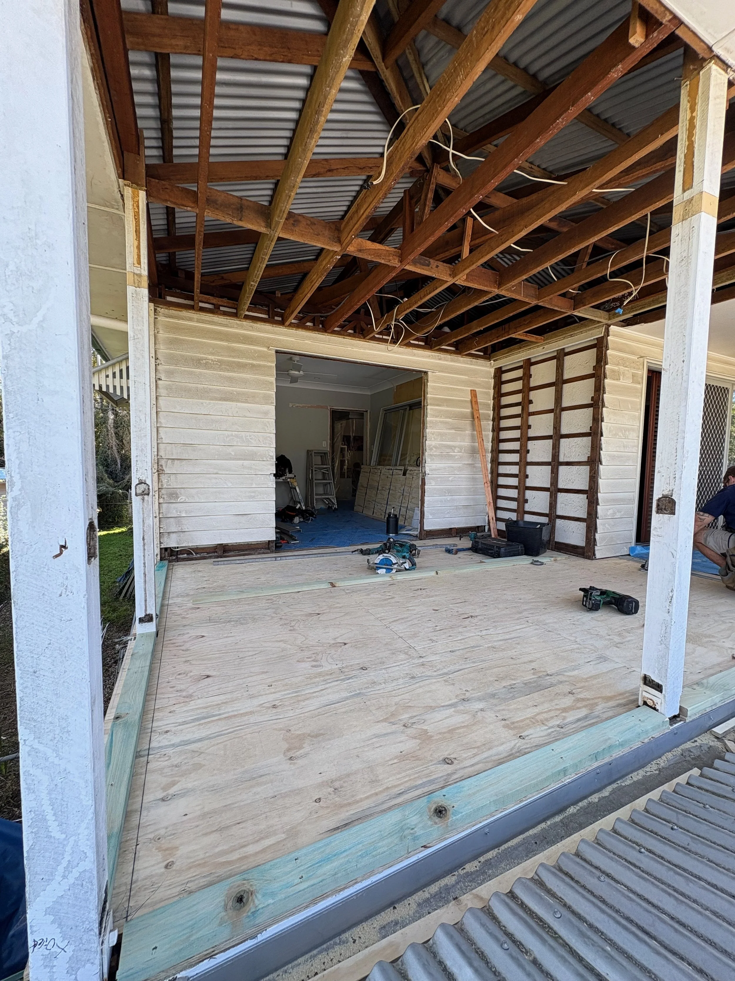 Construction site of a house porch with plywood flooring, exposed wooden framing, and tools such as a drill and saw nearby. The exterior wall has white siding, and the interior room is partially visible through an open doorway.