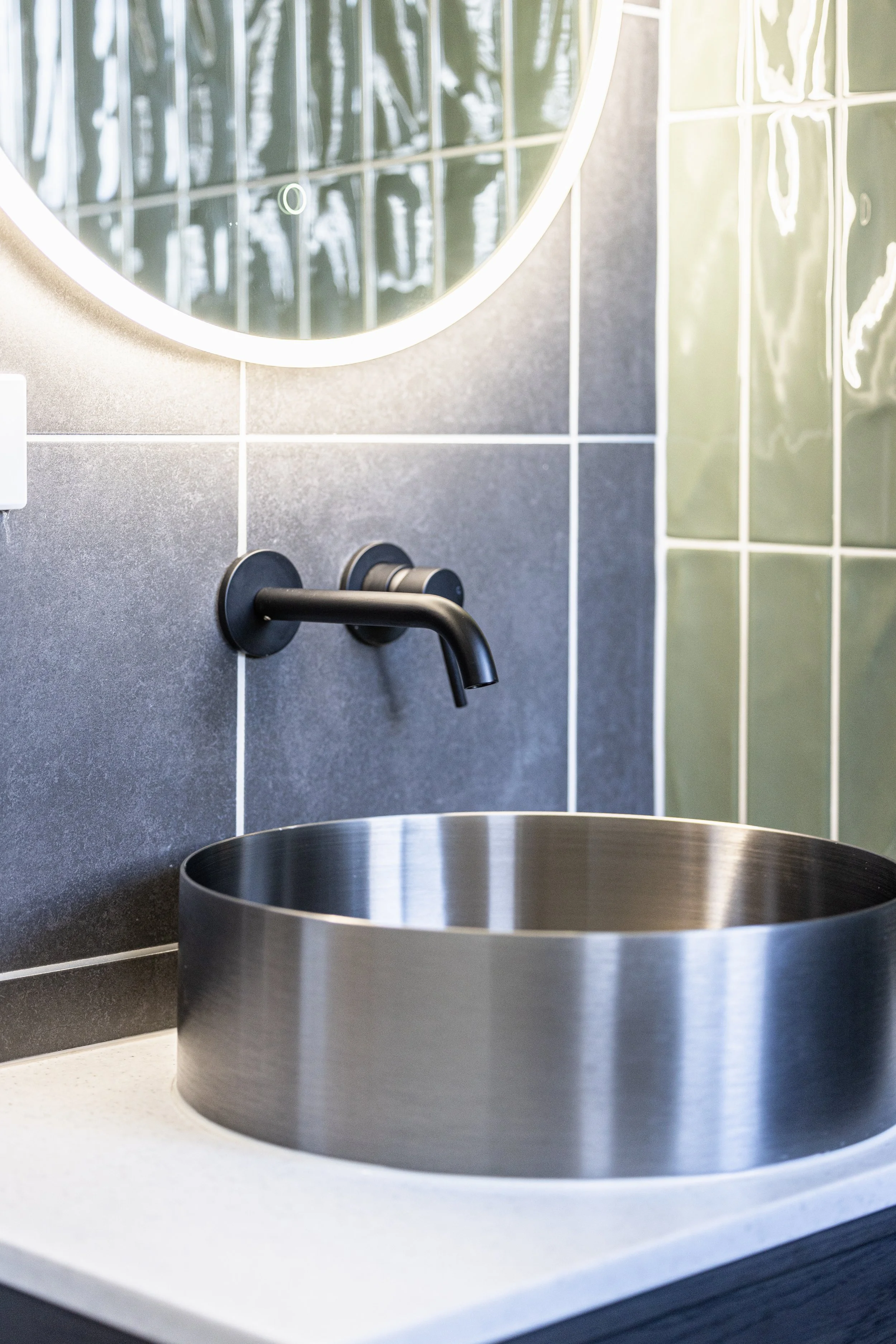 A modern bathroom with a black wall-mounted faucet above a round stainless steel sink and a large round mirror with backlit lighting.