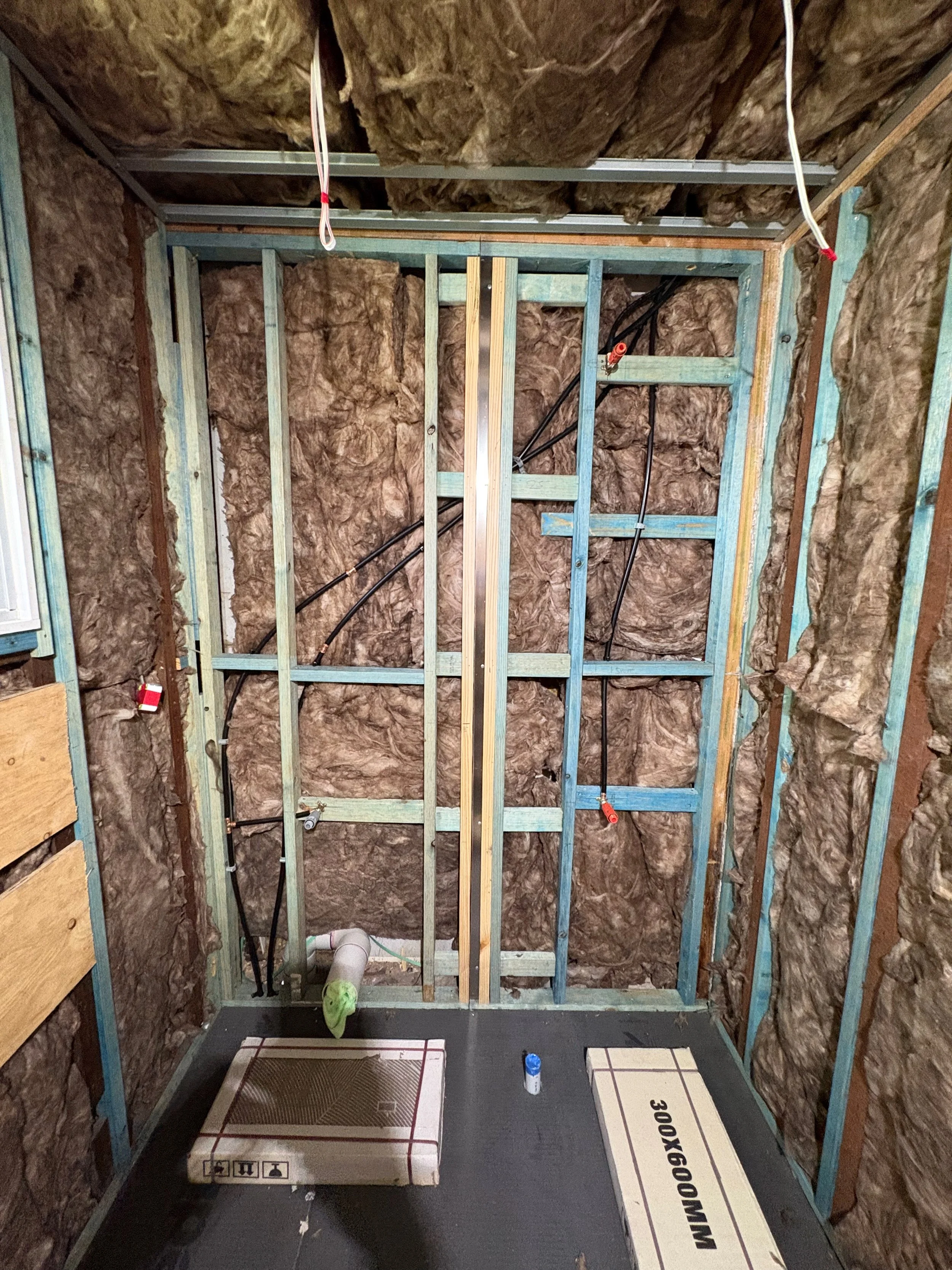 Framing for a bathroom or room under construction with insulation, plumbing pipes, and electrical wiring in the walls.