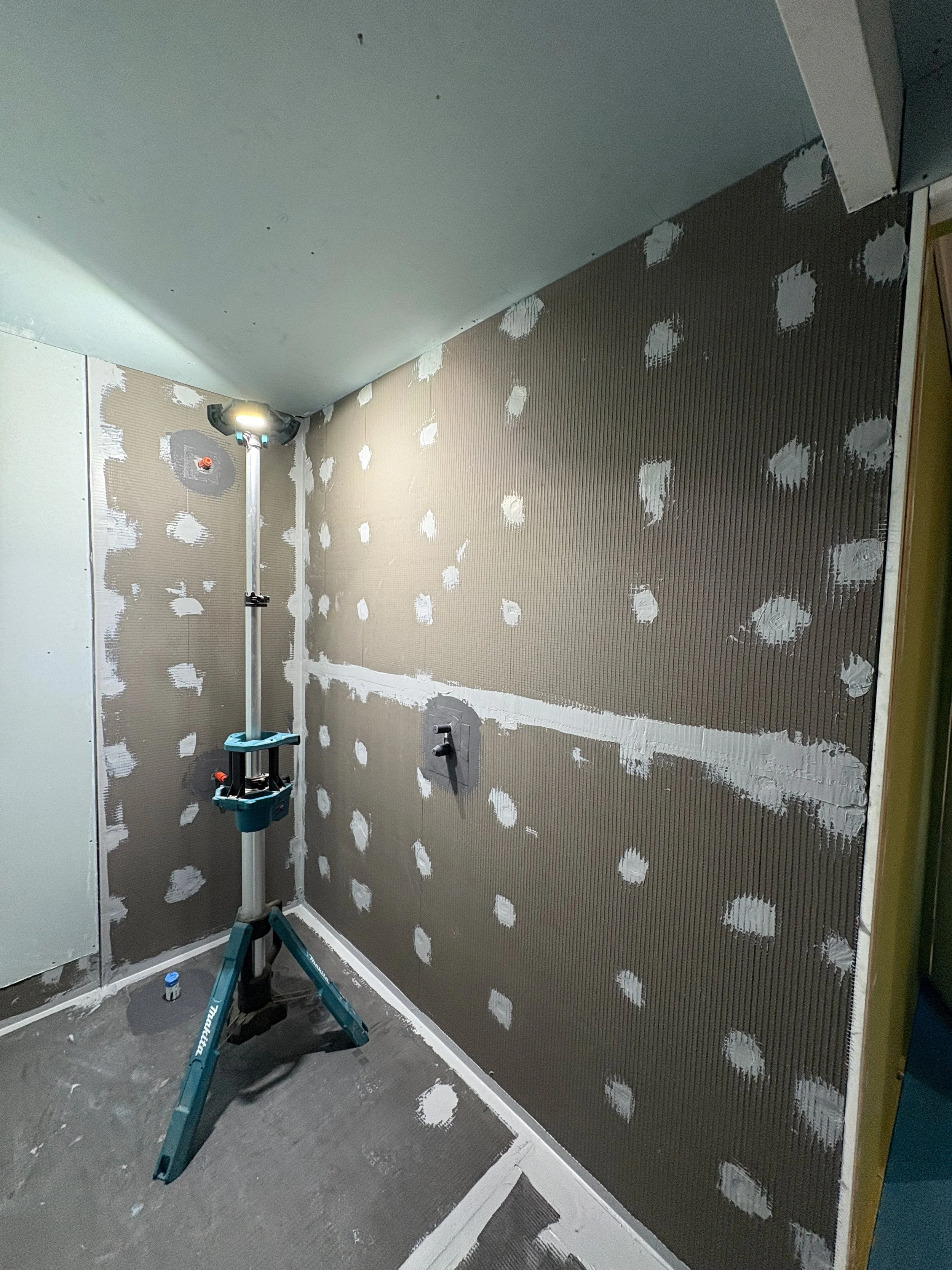 A drywall installation with joint compound on the walls, and a work light on a tripod stand illuminating the space.