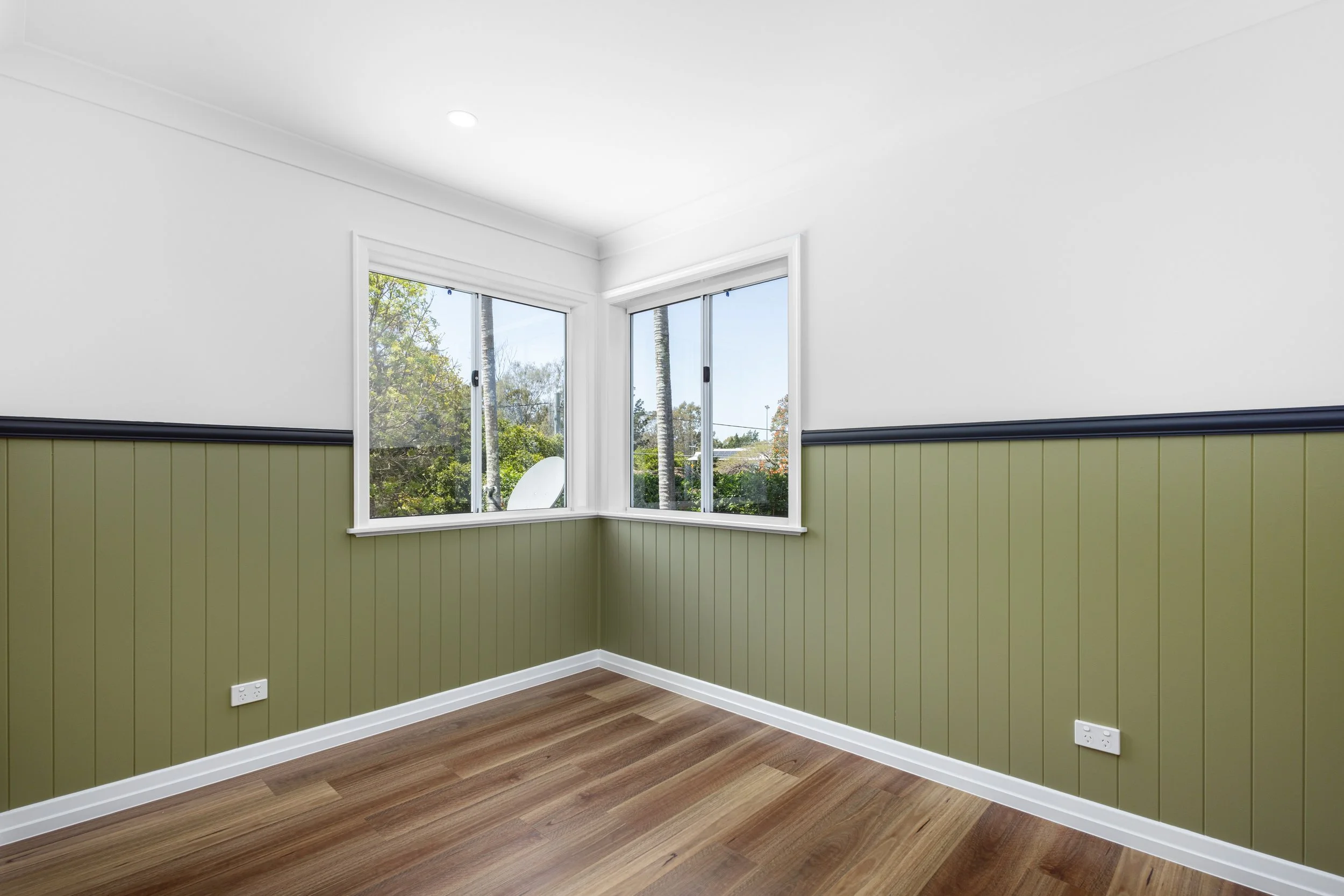 Empty room with white ceiling, white walls, green wainscoting, hardwood floor, and two large windows showing trees outside.