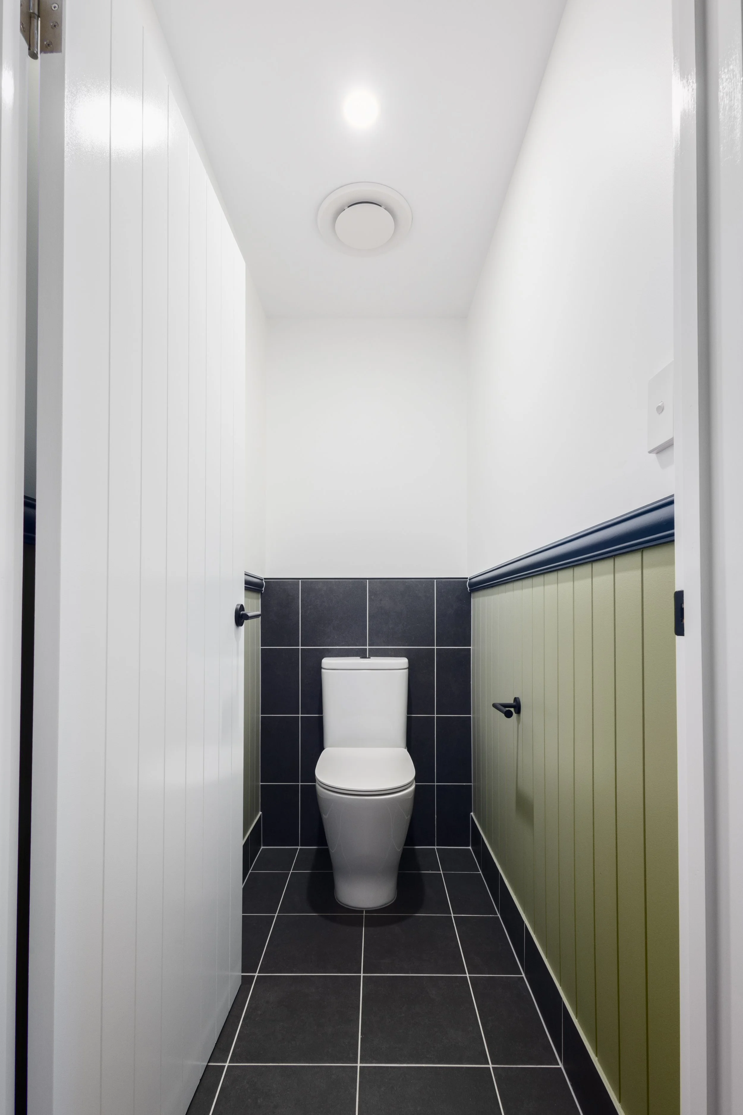 Small modern bathroom with a white toilet, black wall tiles behind it, black and beige wainscoting on the sides, white walls, ceiling lights, and a vent on the ceiling.