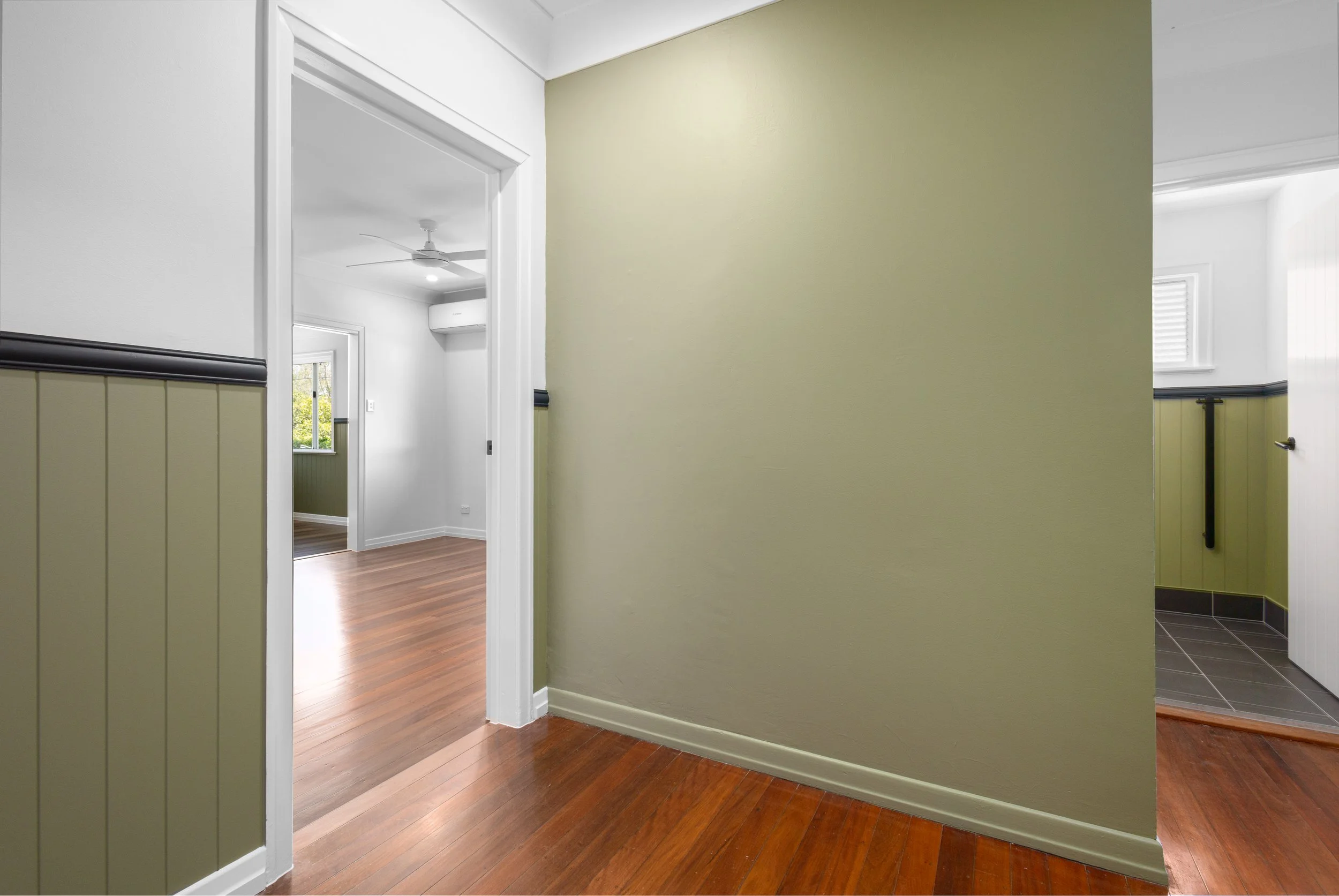 Interior view of a house with hardwood flooring, painted walls in green and white, doorway leading to another room, and a window with a view outside.