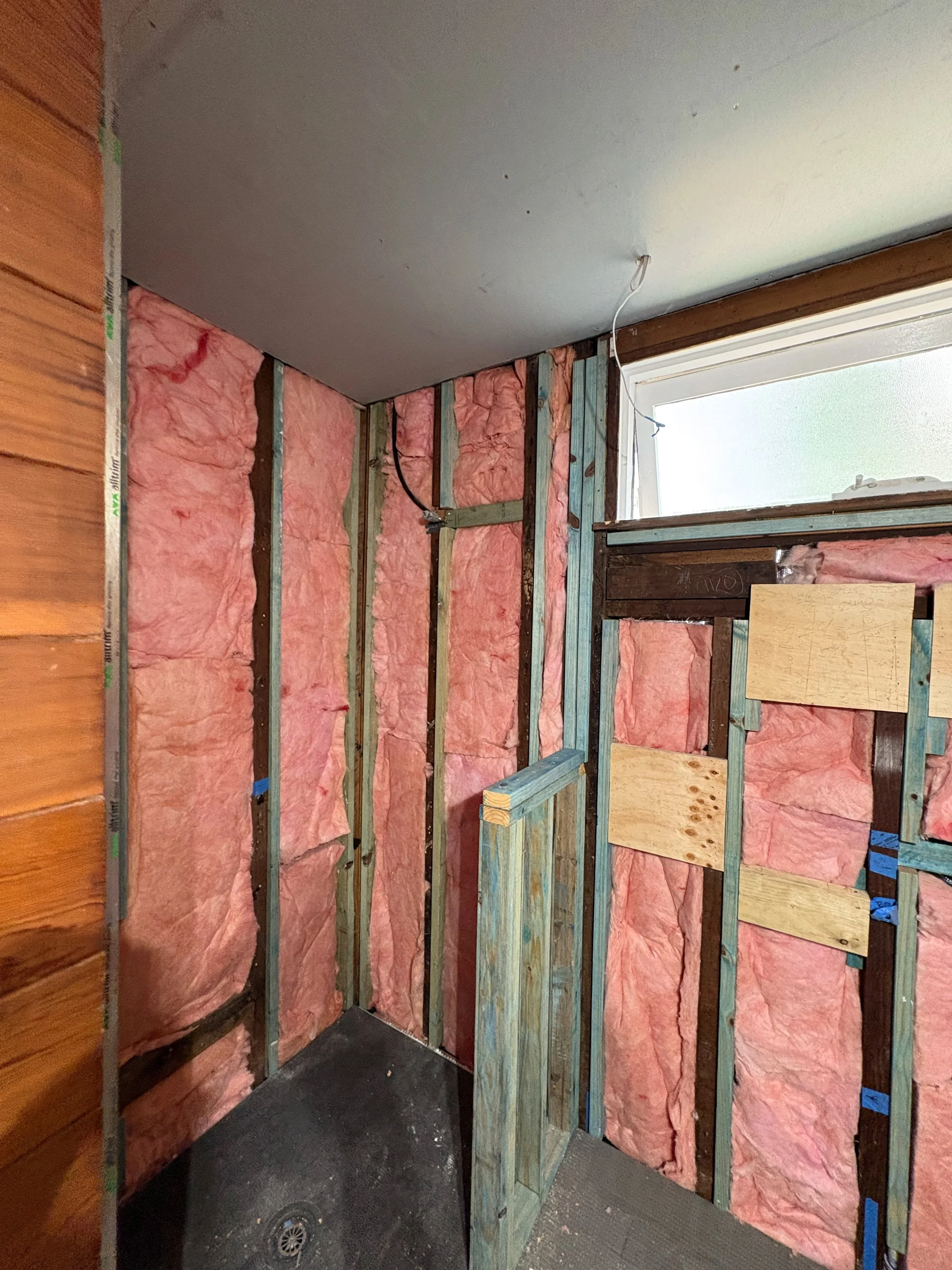 Interior view of a construction site showing pink insulation between wooden studs, with drywall partially installed on the ceiling and some plywood panels attached to the wall.