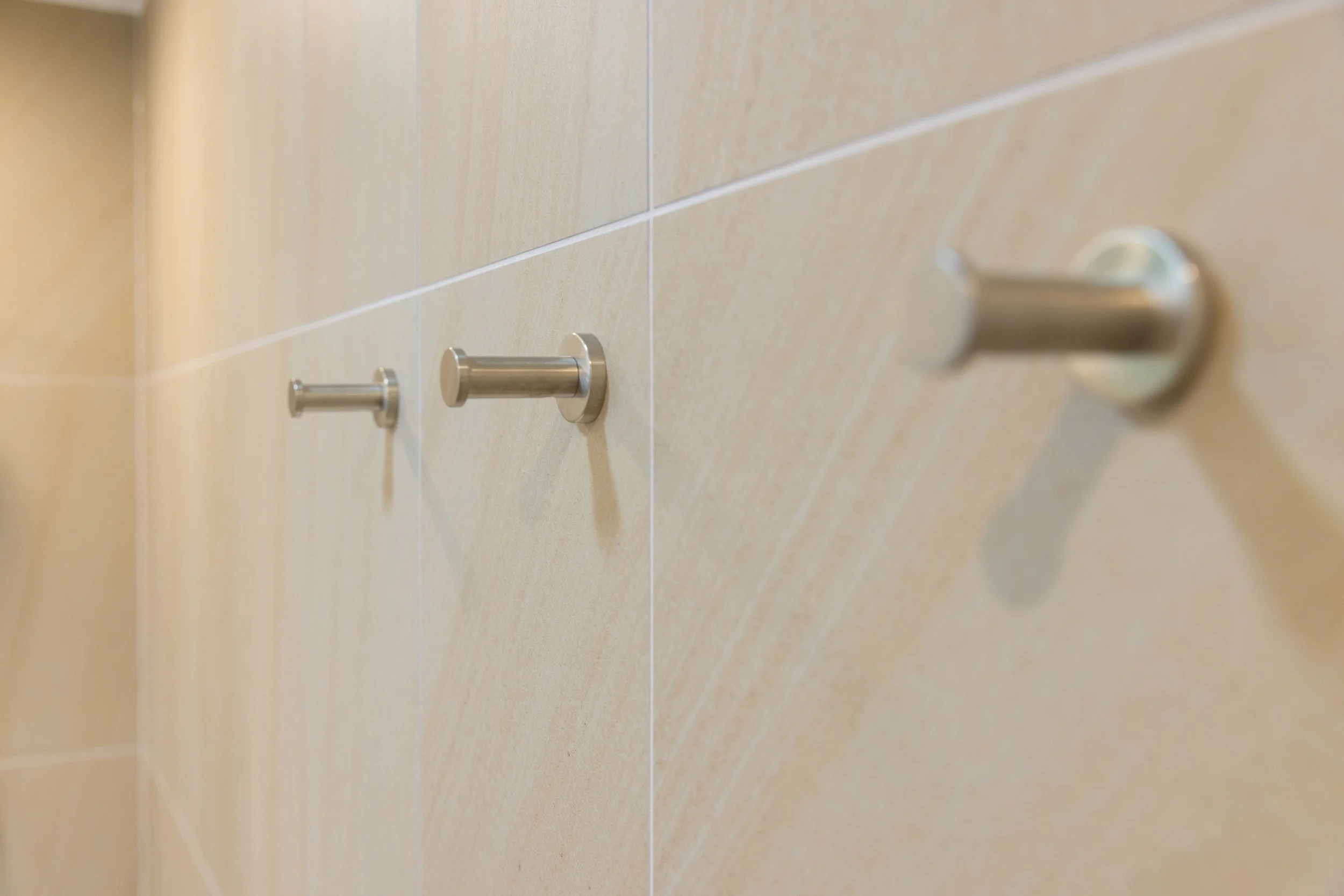 Close-up of three metal hooks mounted on a beige tiled wall.