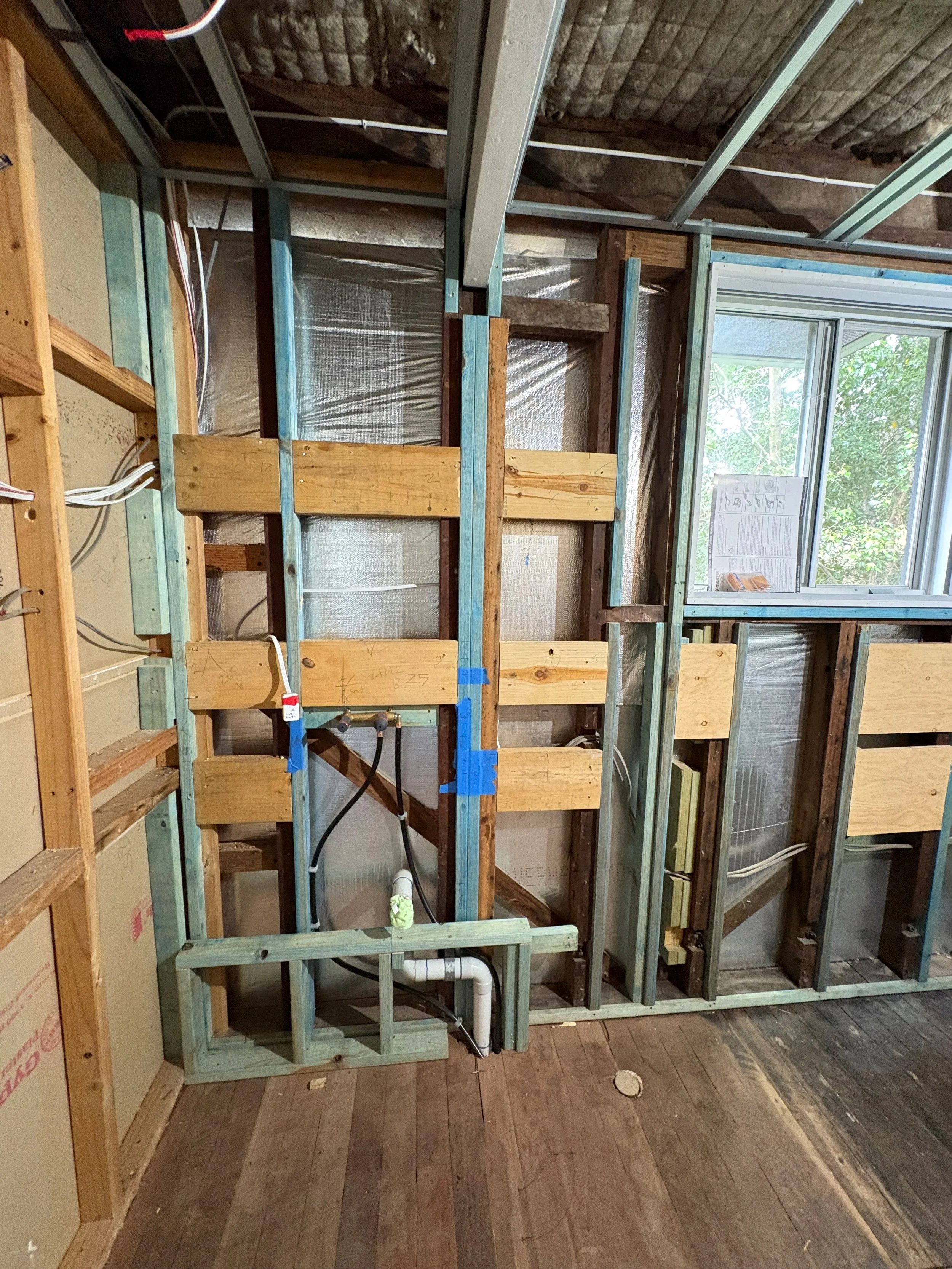 Indoor construction site with wooden framing, electrical wiring, and plumbing pipes, near a window with green foliage outside.