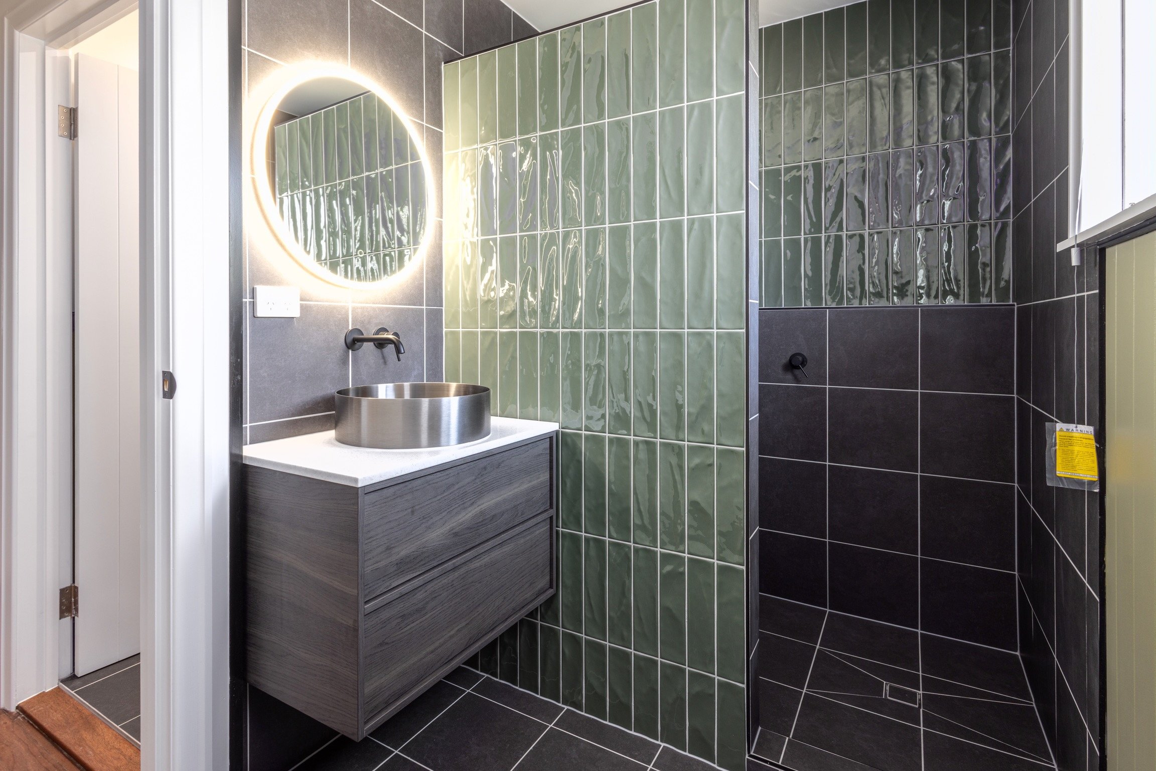 Modern bathroom with black and green tiled walls, a circular mirror with backlighting, a wood vanity with a round stainless steel sink, and a walk-in shower with black tiles.