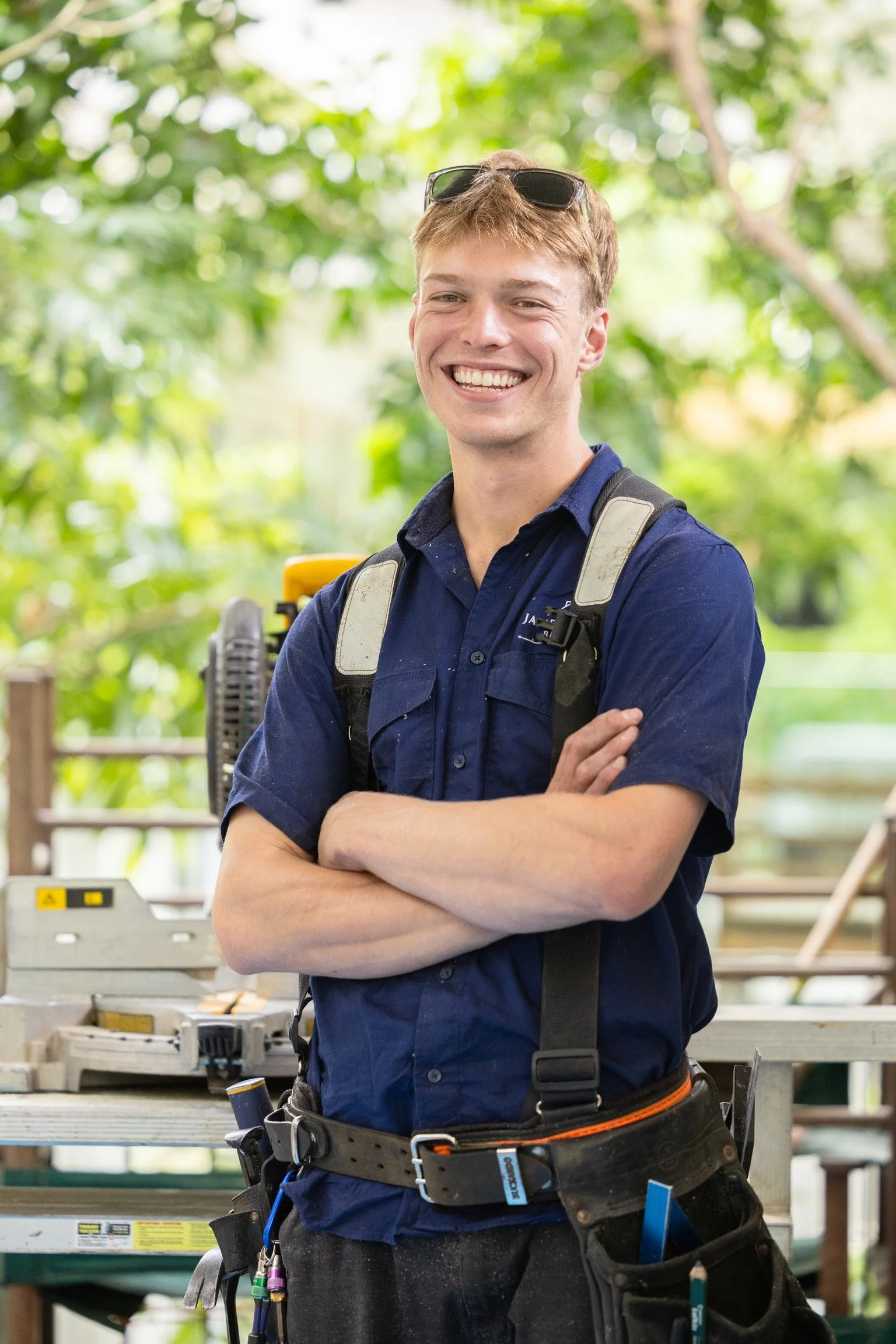 A young man in a navy shirt with sunglasses on his head, smiling with arms crossed, standing outdoors in a lush green environment with tools and equipment around.