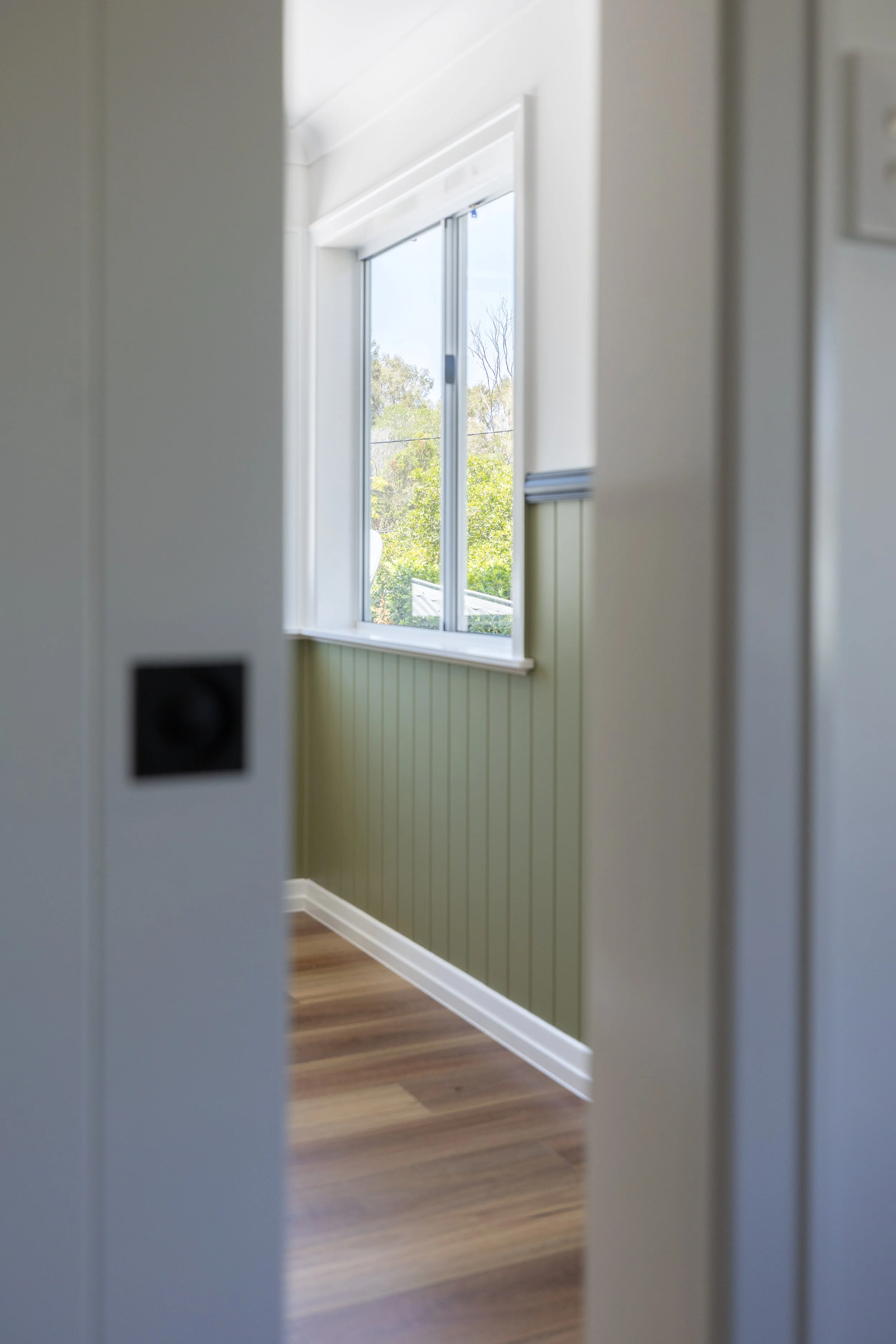 View of a room through a slightly open door, showing a window with a view of trees outside, beige wall paneling below the window, and hardwood flooring.