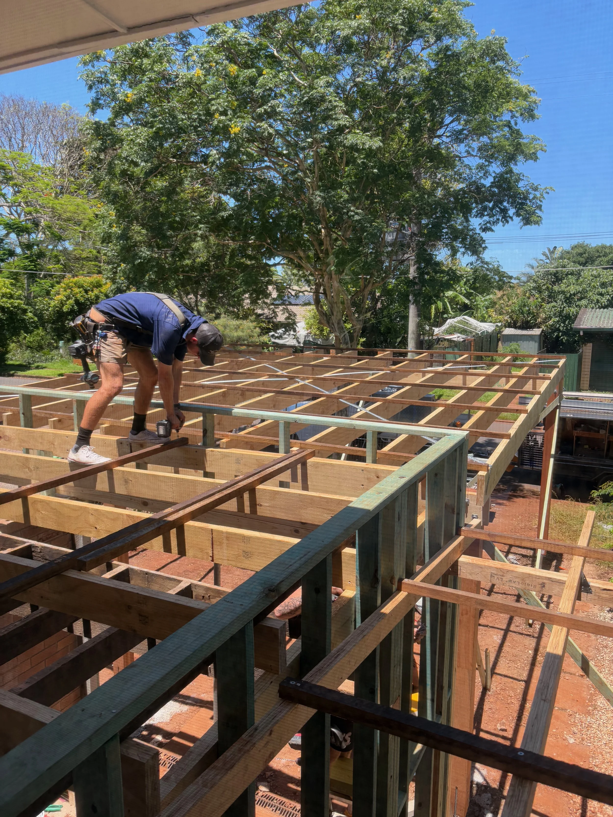 A person working on a wooden construction site, using a power drill, with a large tree and clear blue sky in the background.