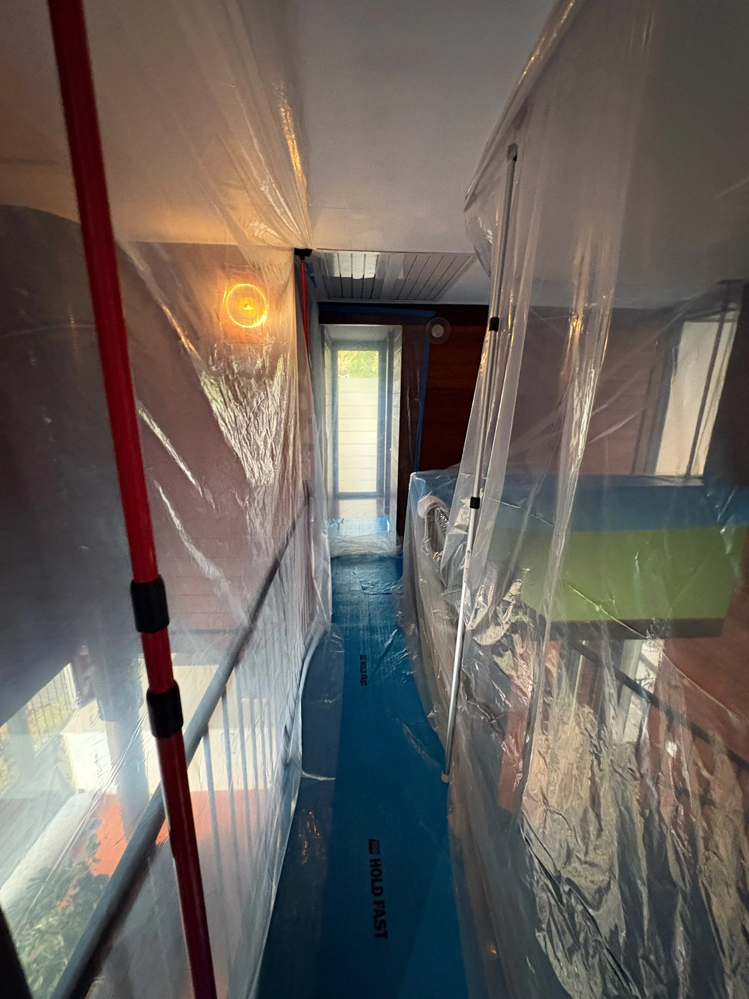 A narrow, plastic-covered passageway inside a construction or renovation site, with a window at the end letting in natural light. The floor is covered with blue protective material labeled "HOLD FAST," and there is a red pole with black tape on the l