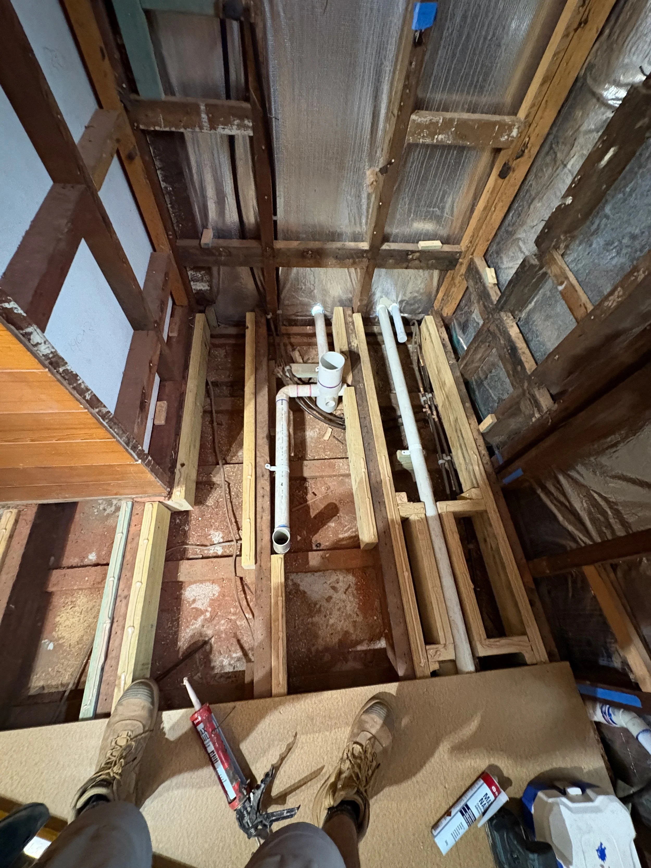 View of a bathroom under construction with exposed wooden framing and plumbing pipes, seen from a person's perspective standing on a raised platform.