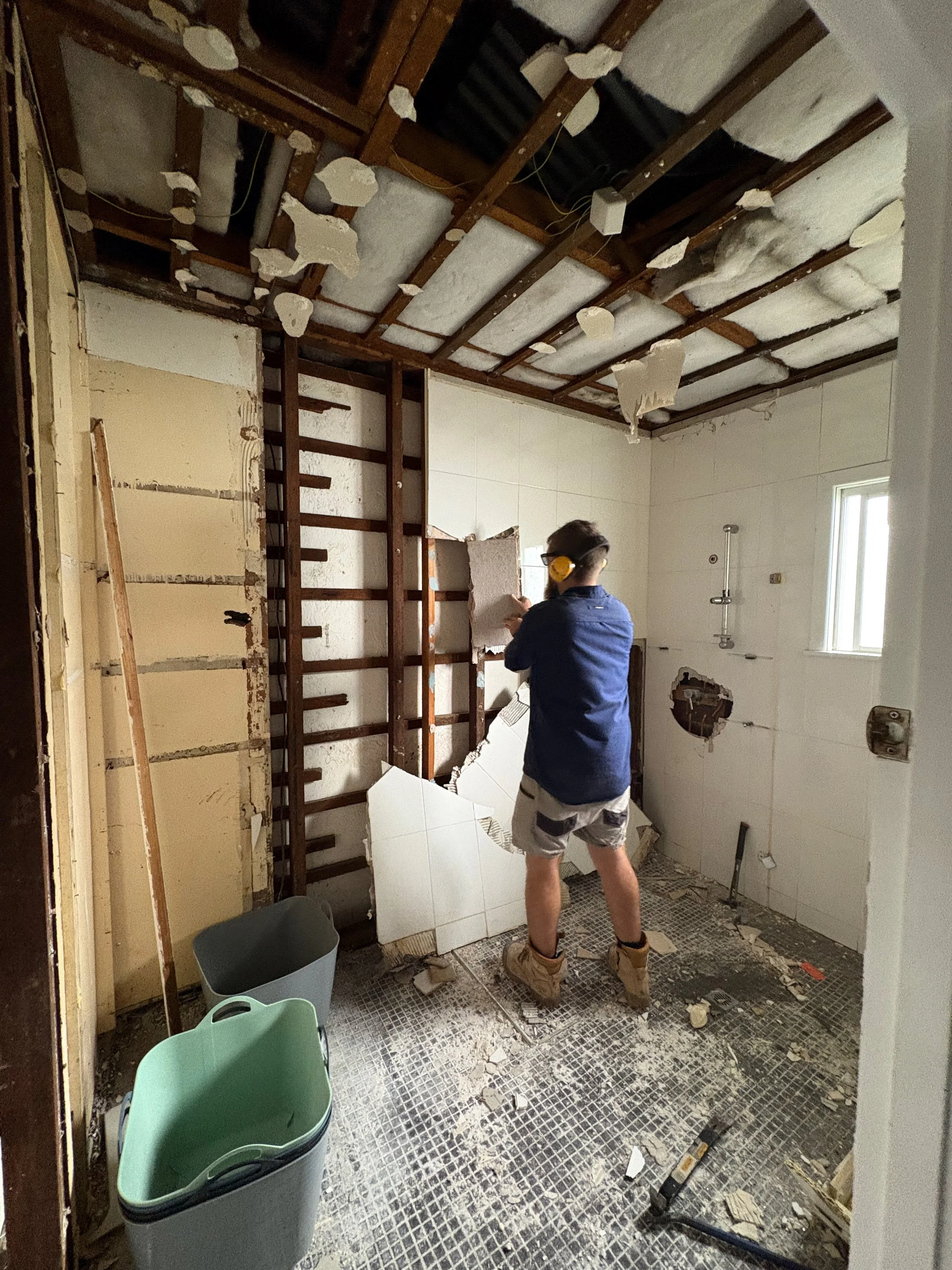 A person wearing protective ear muffs and work boots demolishing a bathroom wall with a hammer and chisel, with exposed ceiling insulation and debris on the floor.