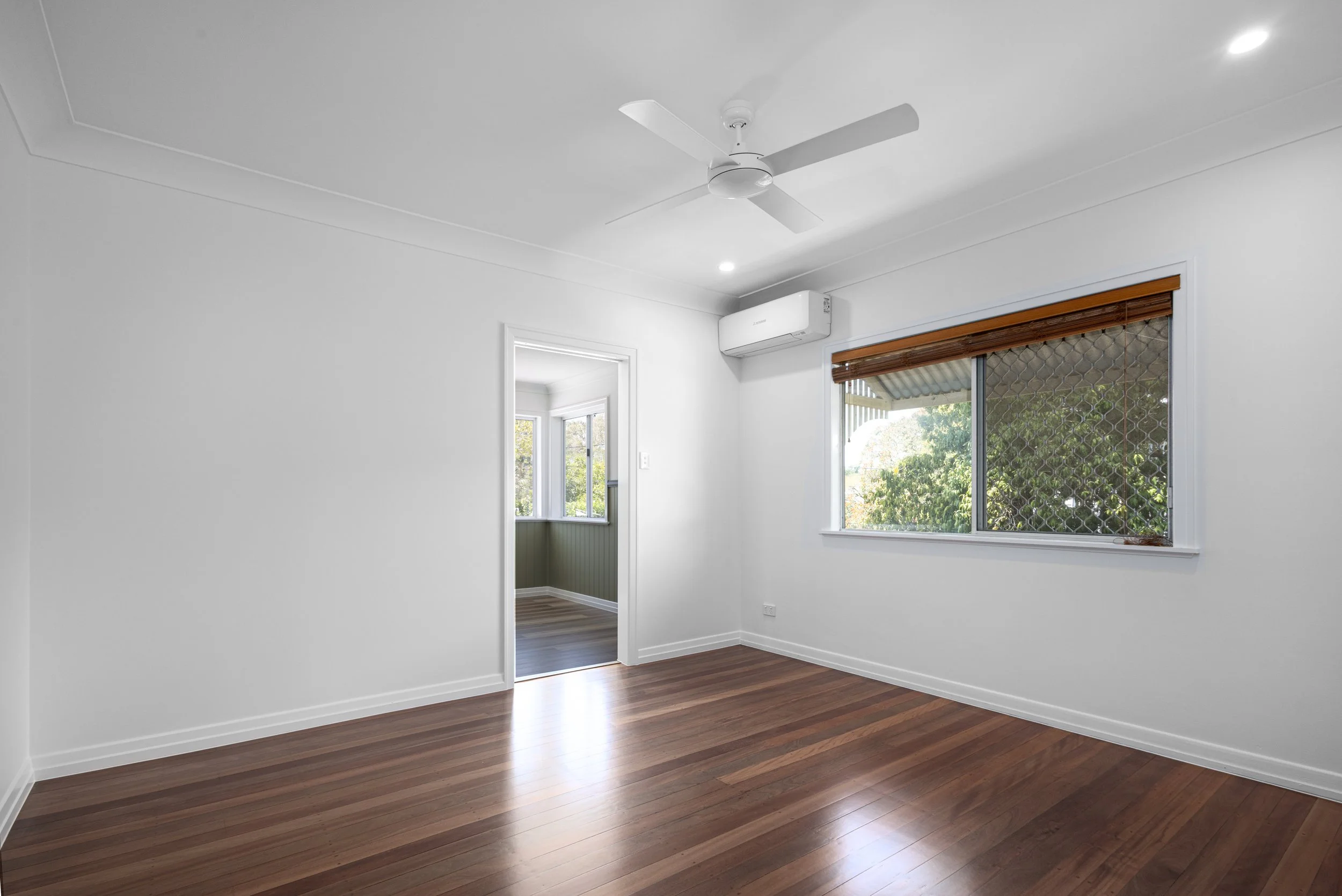 Empty room with white walls, wooden floor, window with brown blinds, ceiling fan, and air conditioning unit.