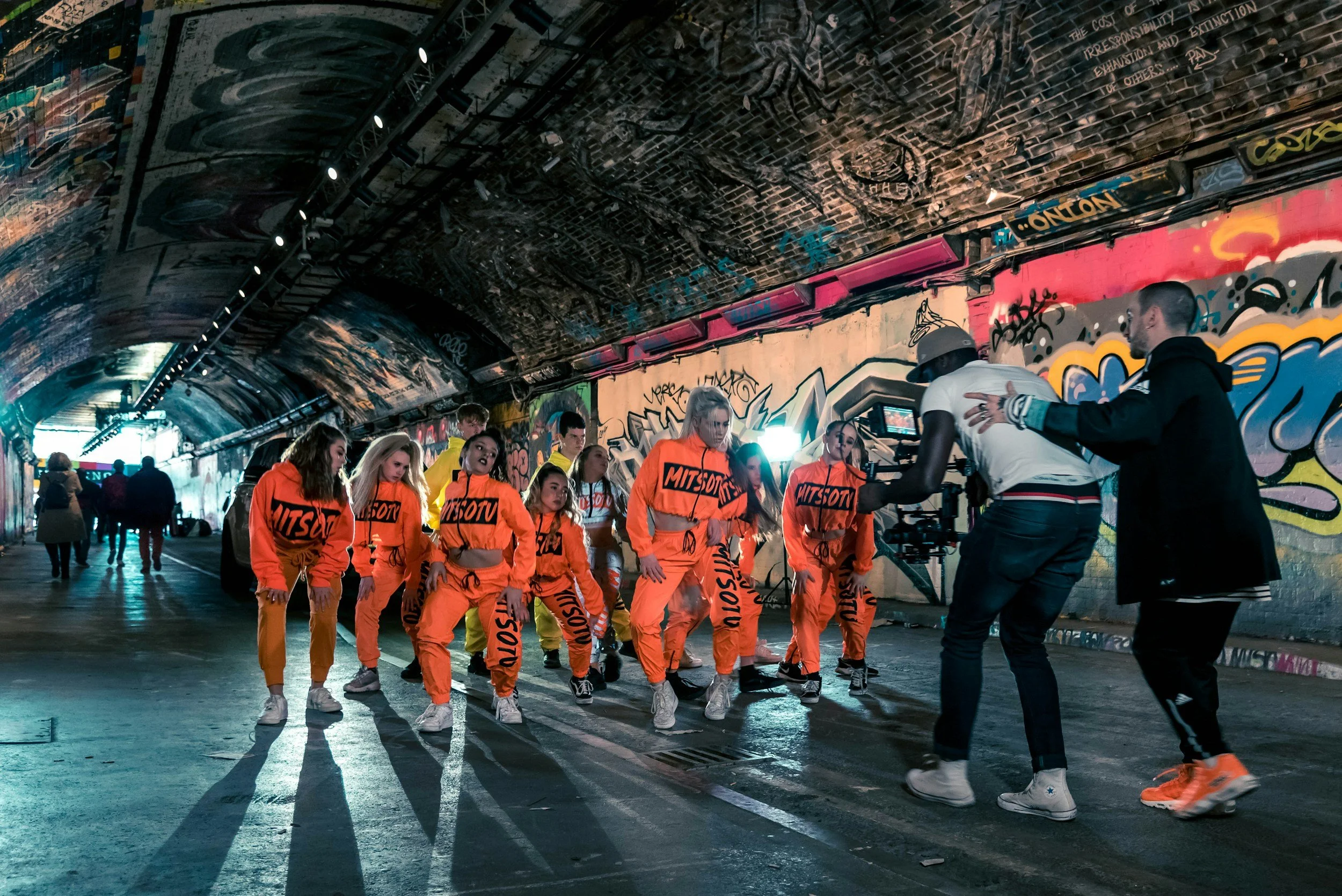 Un gruppo di ballerini in abbigliamento arancione si esibiscono in un tunnel graffiti, con due persone che riprendono la scena con una videocamera.