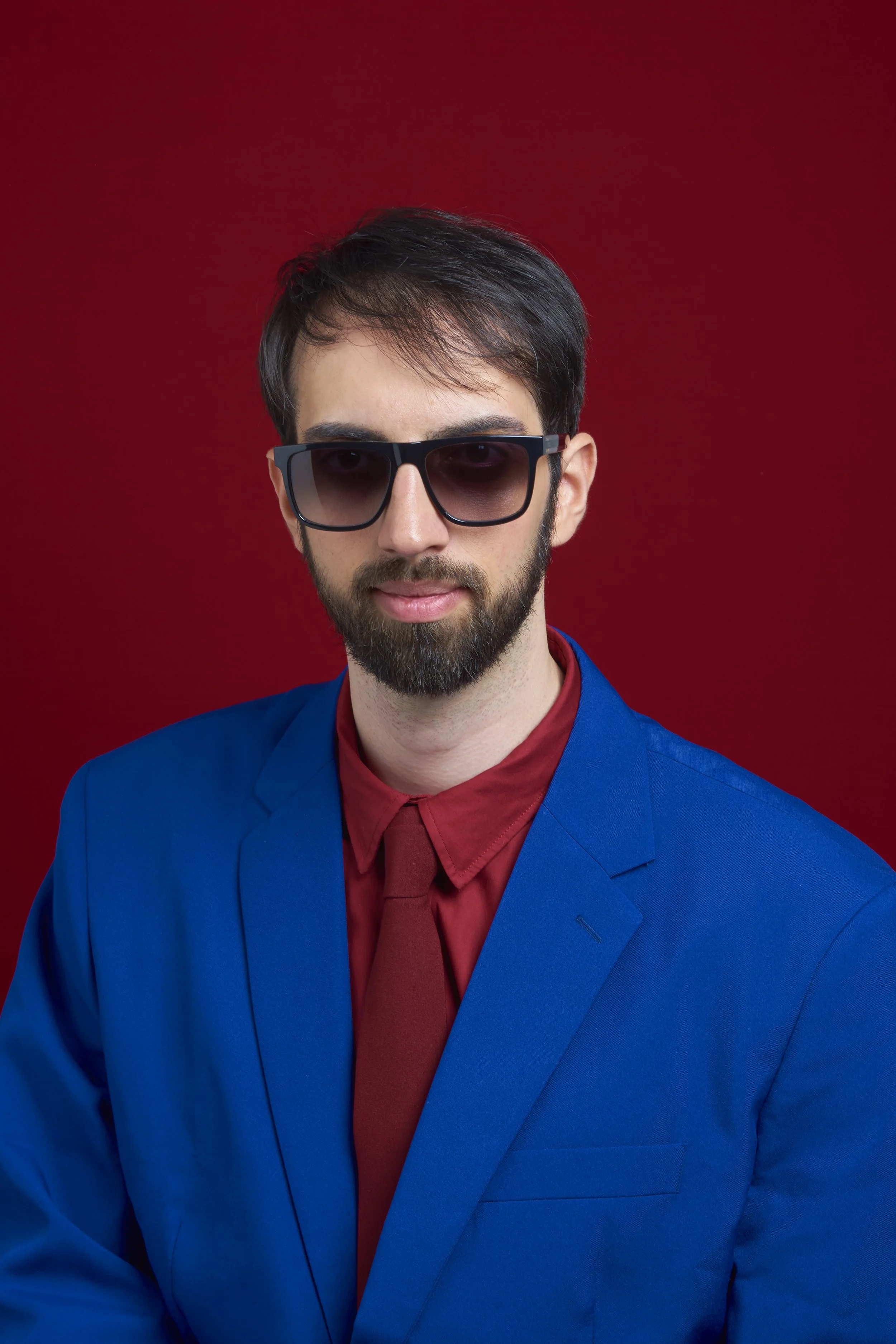 Ritratto di un uomo con barba, occhiali da sole scuri, abito blu, camicia rossa e cravatta rossa, con sfondo rosso.