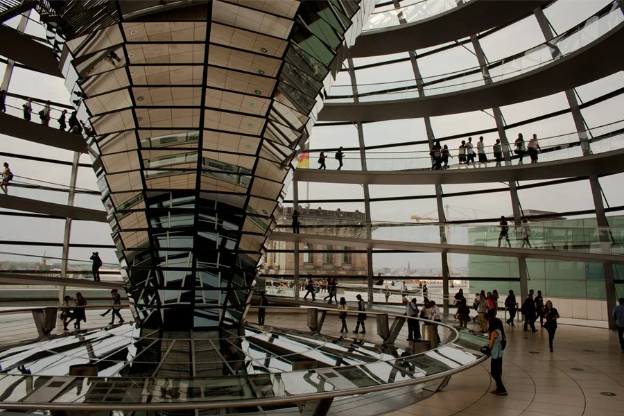 Interior view of the Reichstag Dome in Berlin, Germany, with people walking on spiral ramps around the mirrored central structure.