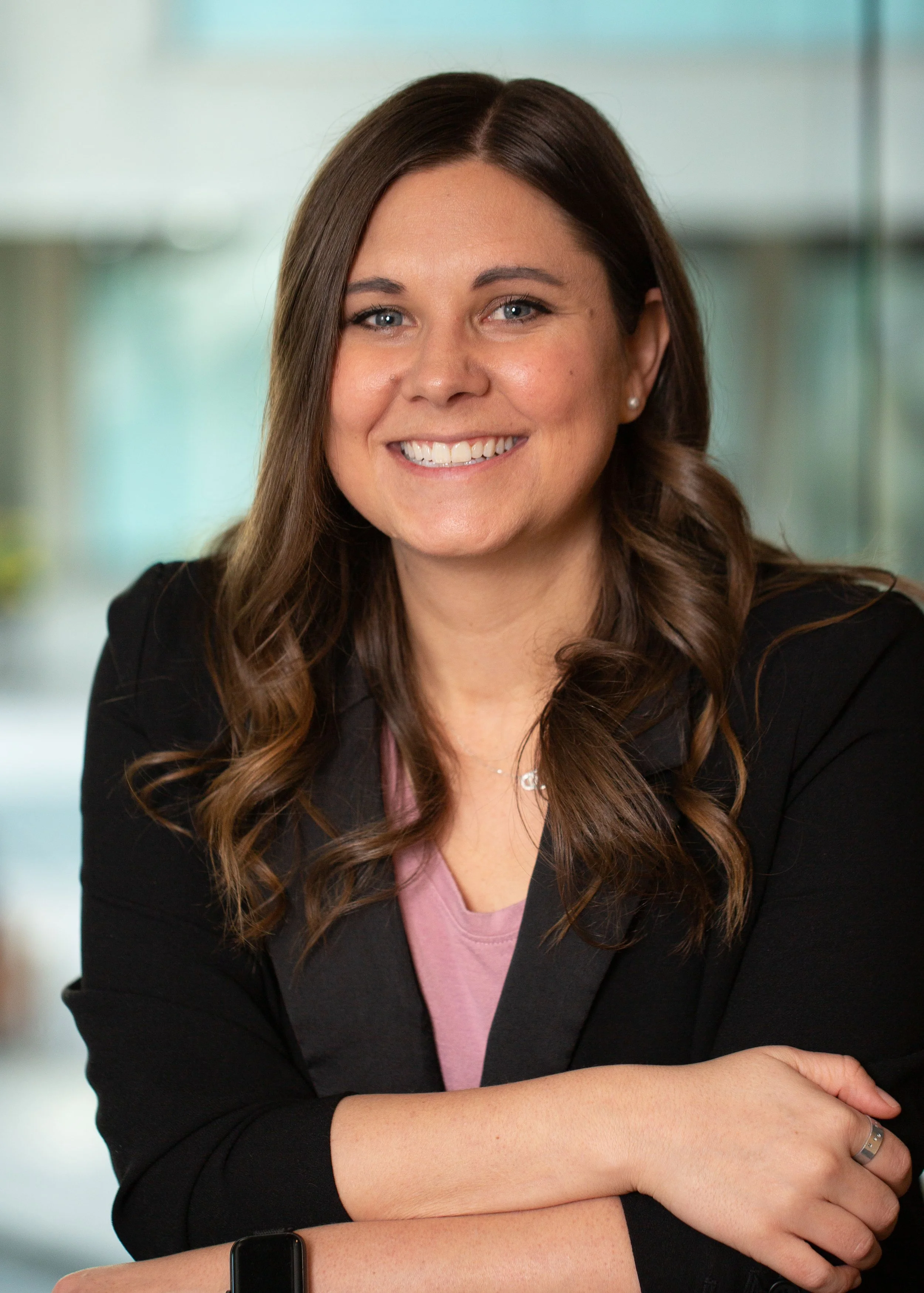 A smiling woman with long brown hair, wearing a black blazer and pink top, with her arms crossed, standing in an office setting.