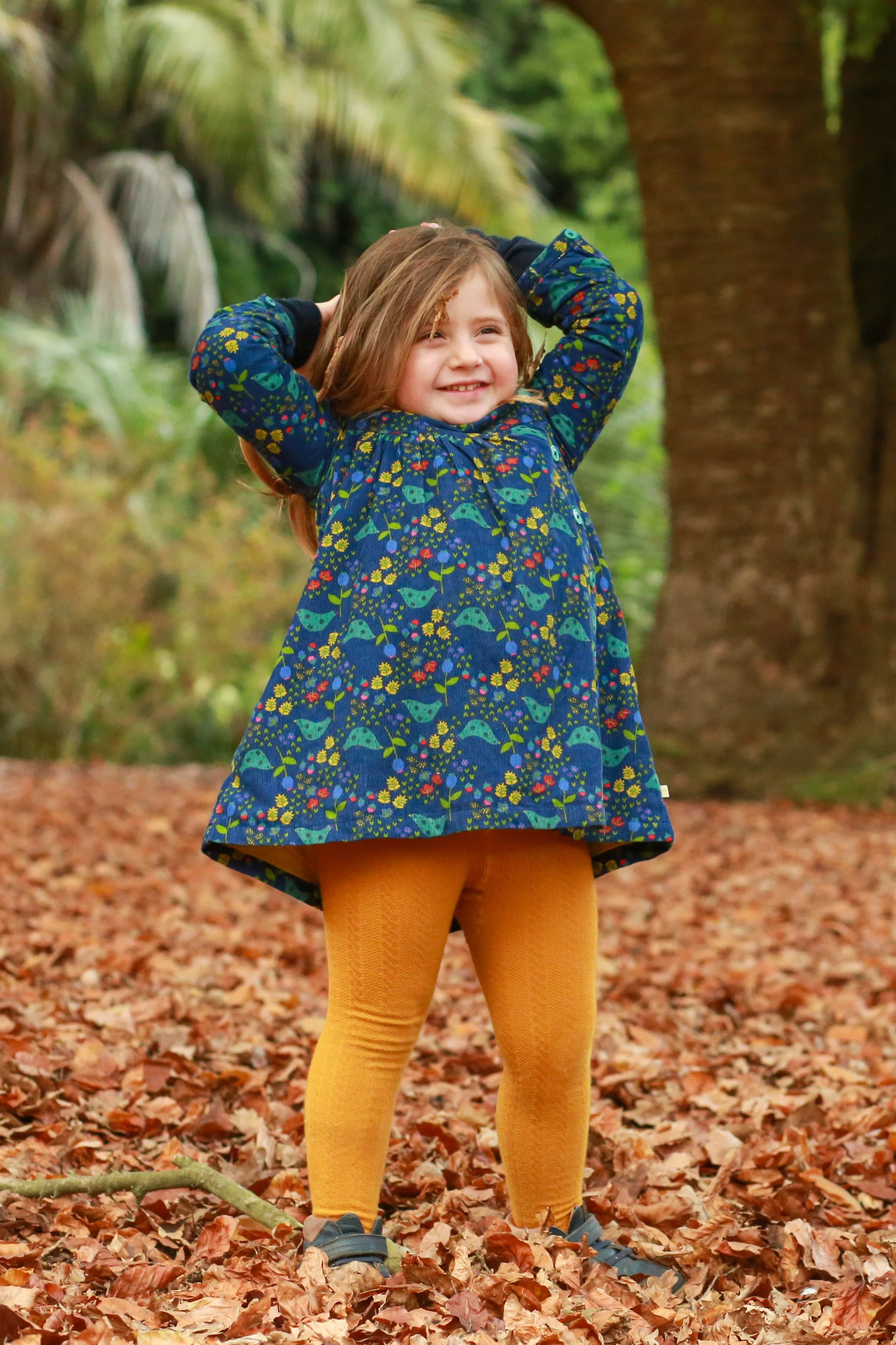 A young girl standing on a leaf-covered ground in a forest, smiling and holding her hands behind her head.
