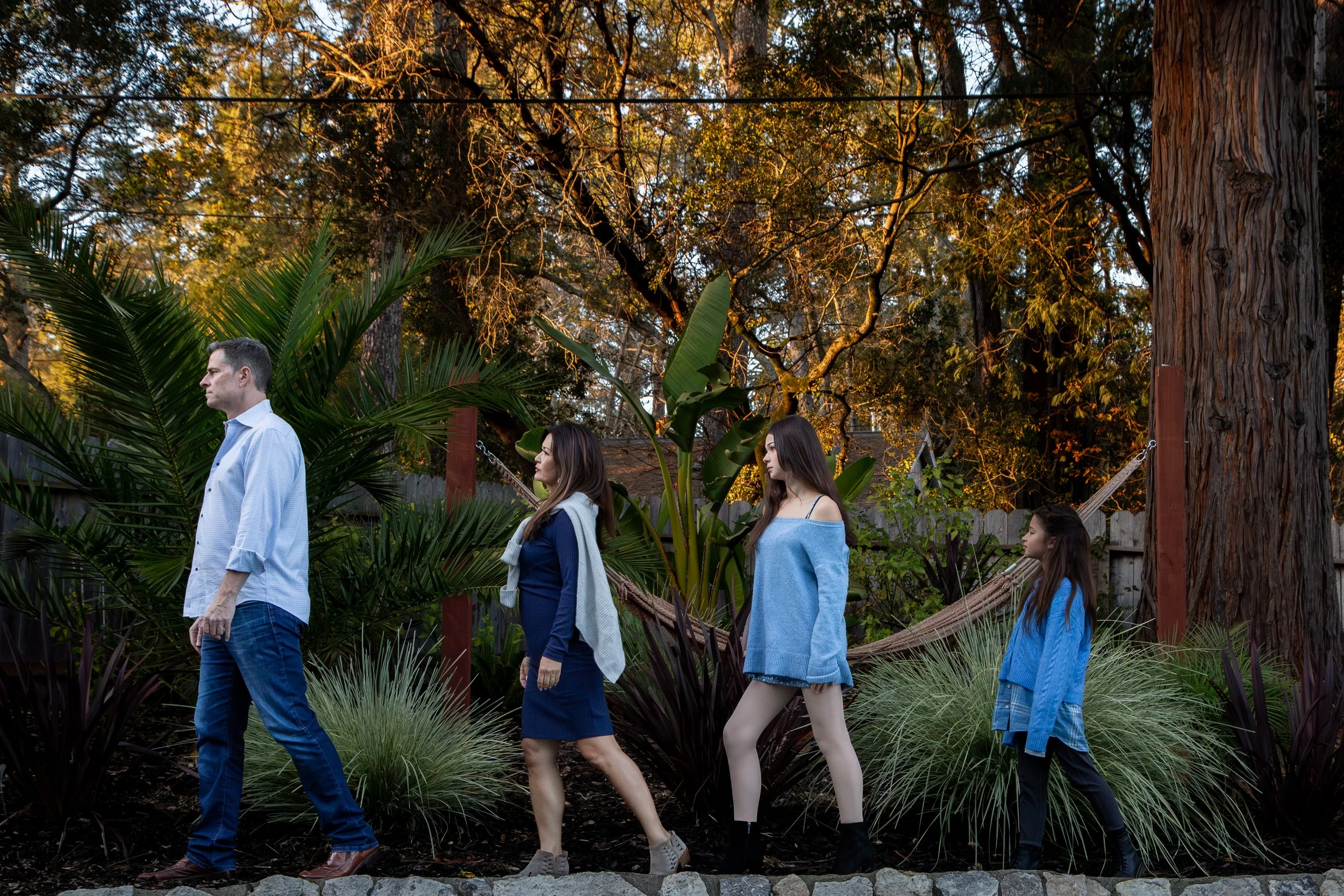 A family walking in a backyard garden at sunset with large trees, bushes, and a hammock.