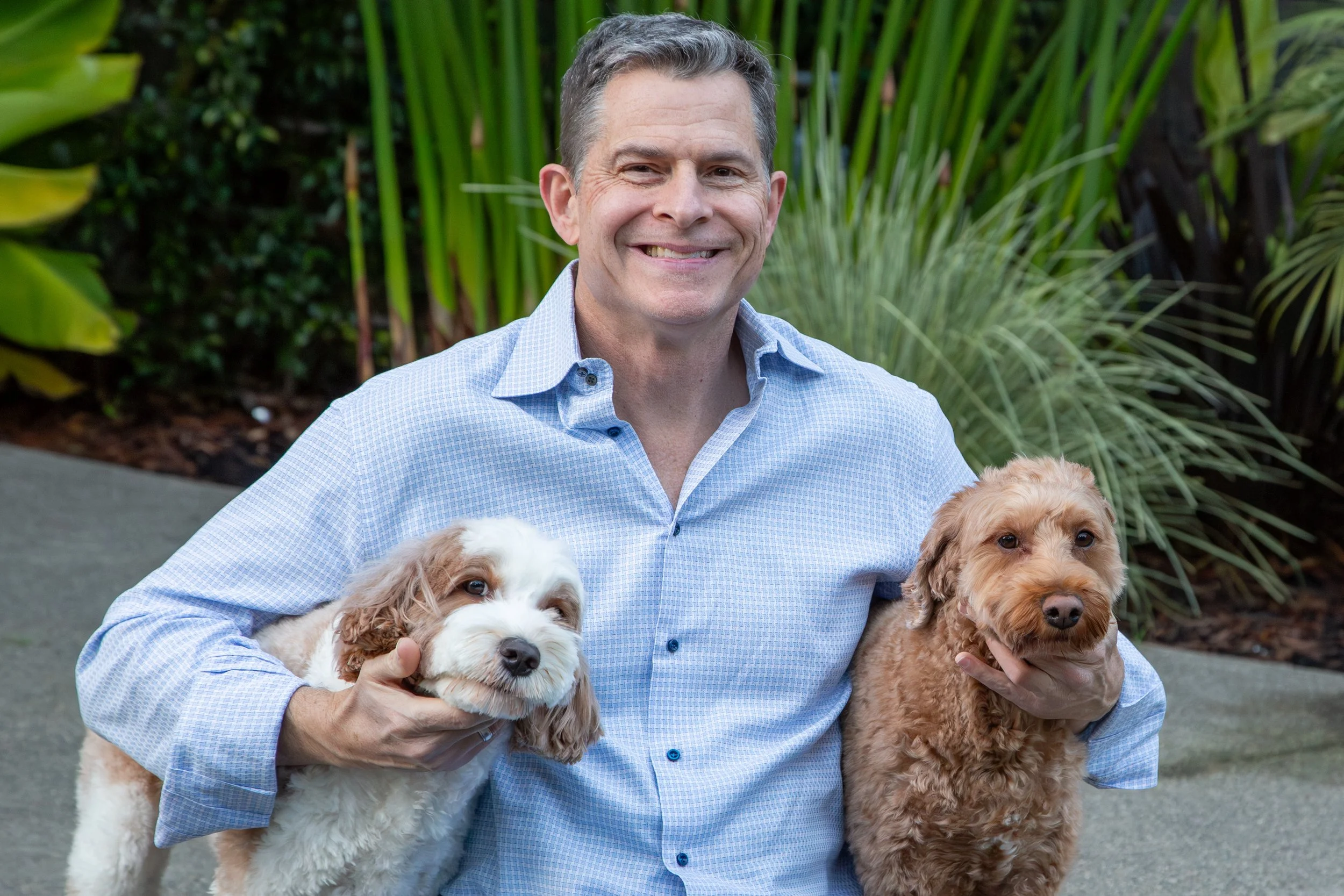 Smiling man in a blue checkered shirt holding two dogs, one white and brown, the other brown, outdoors with greenery in the background.