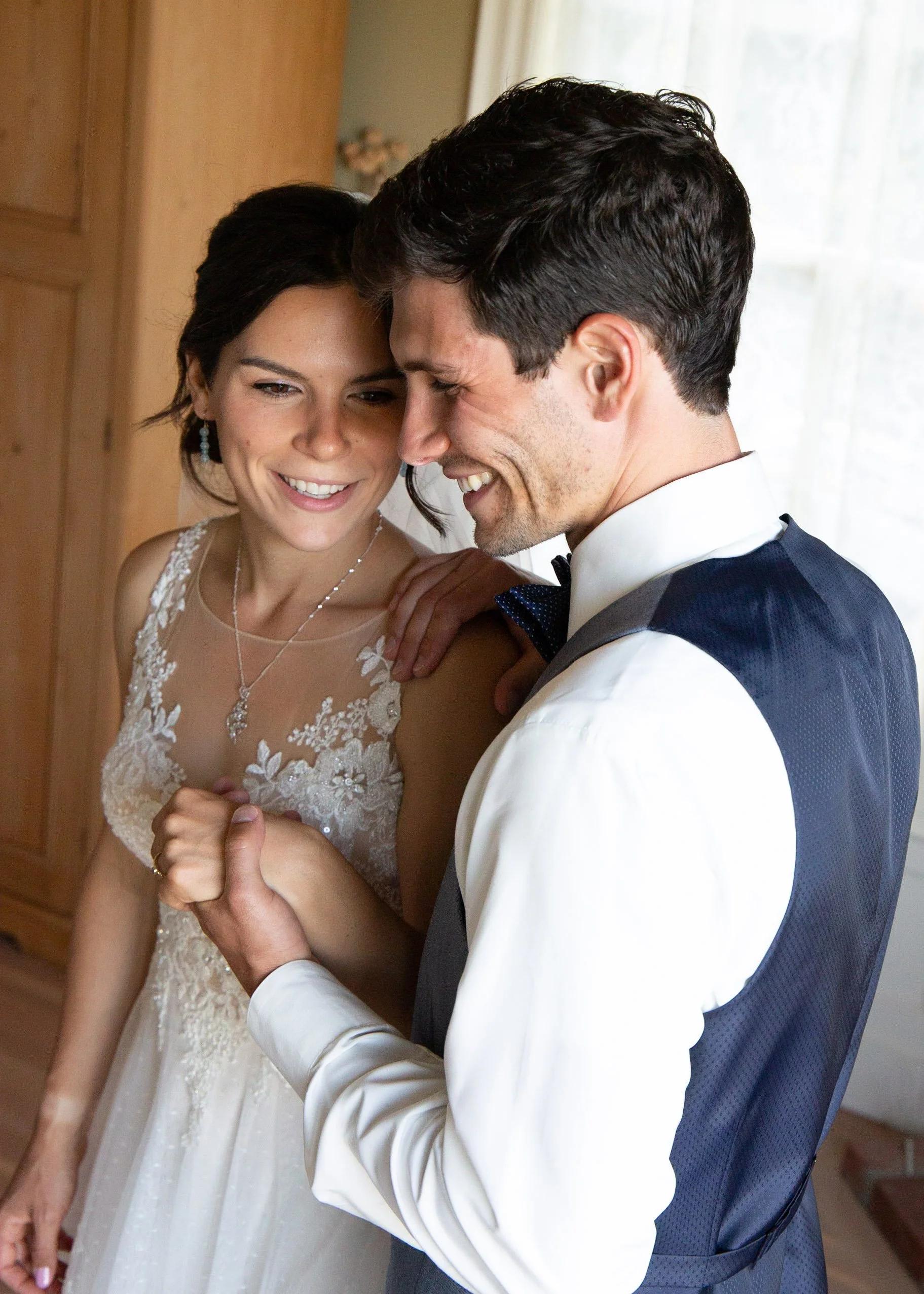 A bride and groom holding hands and smiling, dressed in wedding attire indoors with wooden furniture and a window with curtains in the background.