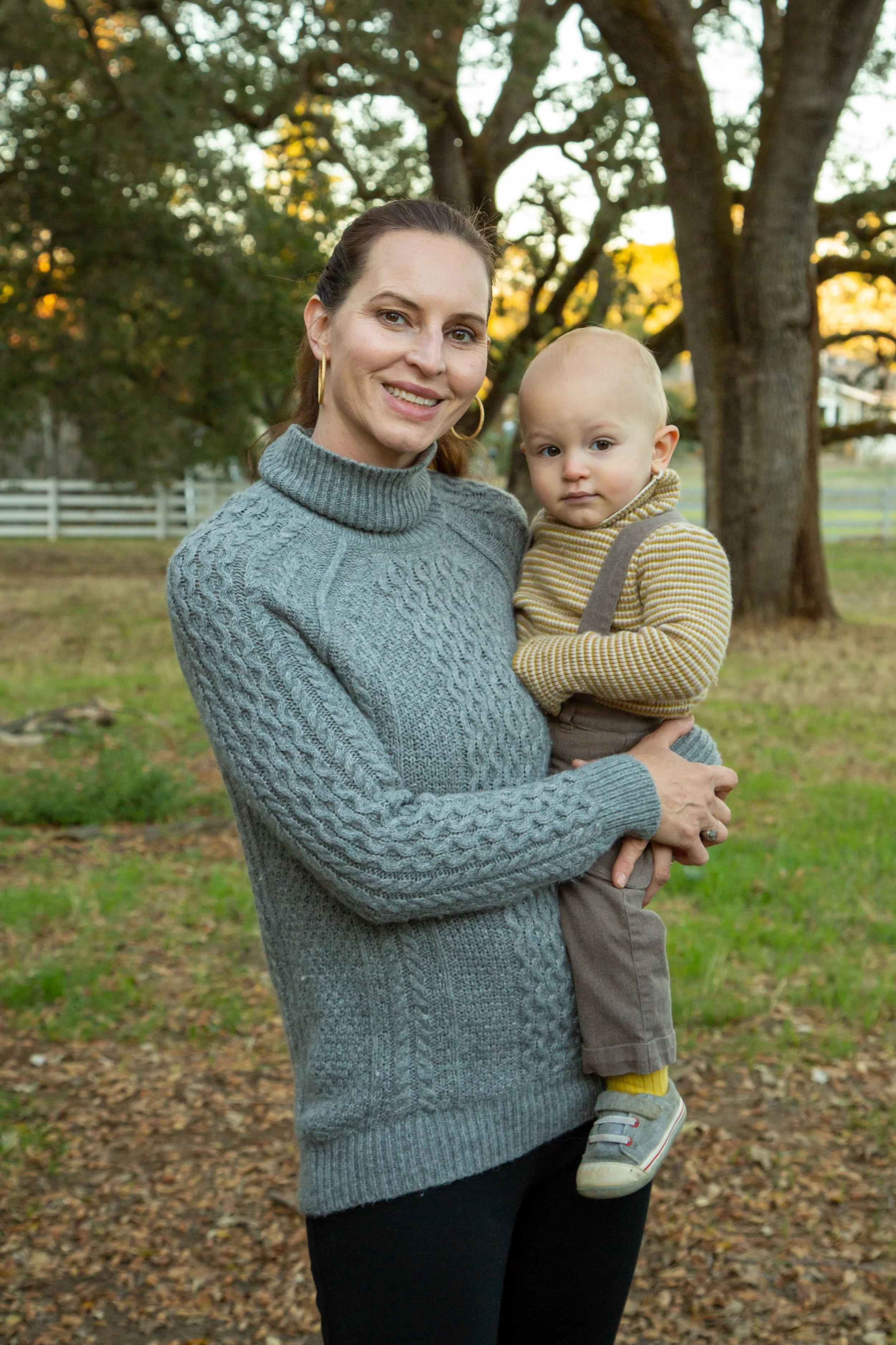 A woman with brown hair in a ponytail, wearing a gray cable-knit turtleneck sweater, holding a young boy with blonde hair, wearing a yellow and brown striped sweater, in a park with green grass and large trees with autumn leaves.
