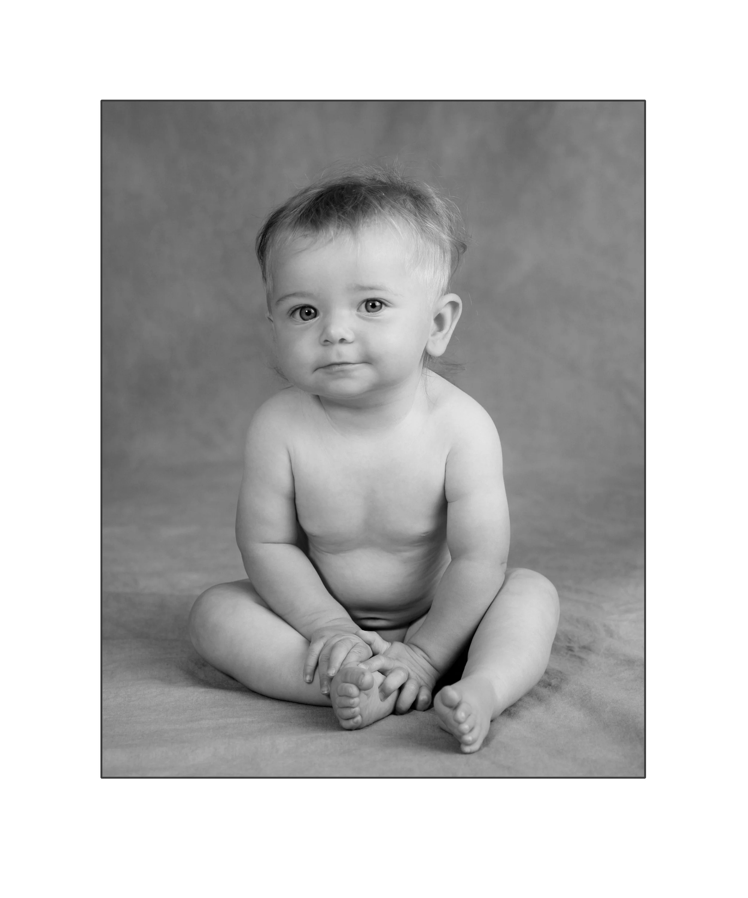 A black and white photo of a young, barefoot baby with light hair sitting on a textured surface, looking directly at the camera.