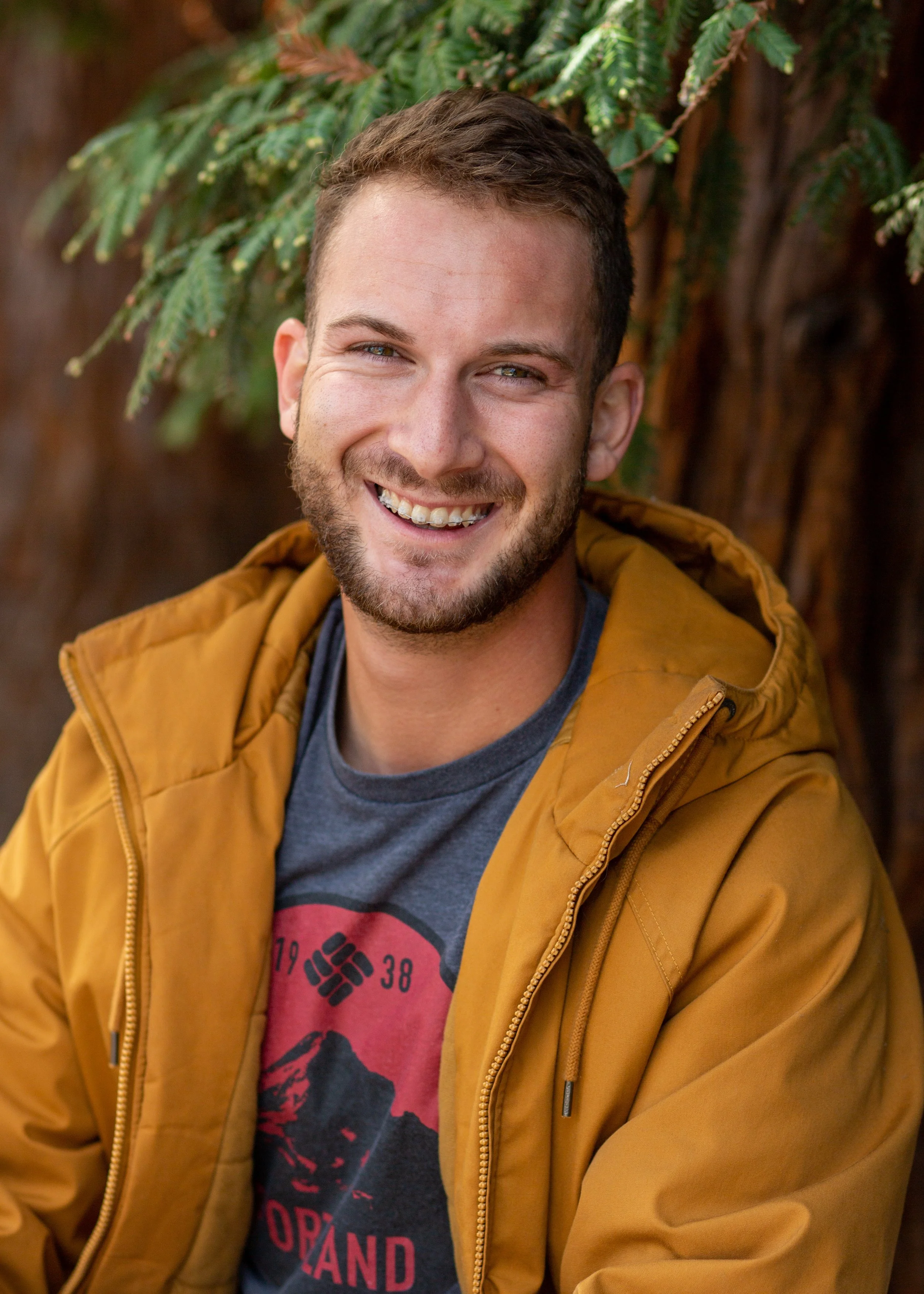 A smiling young man with light brown hair and a beard, wearing a mustard yellow jacket and a gray t-shirt with red and black graphic, standing outdoors with a large tree and green foliage in the background.