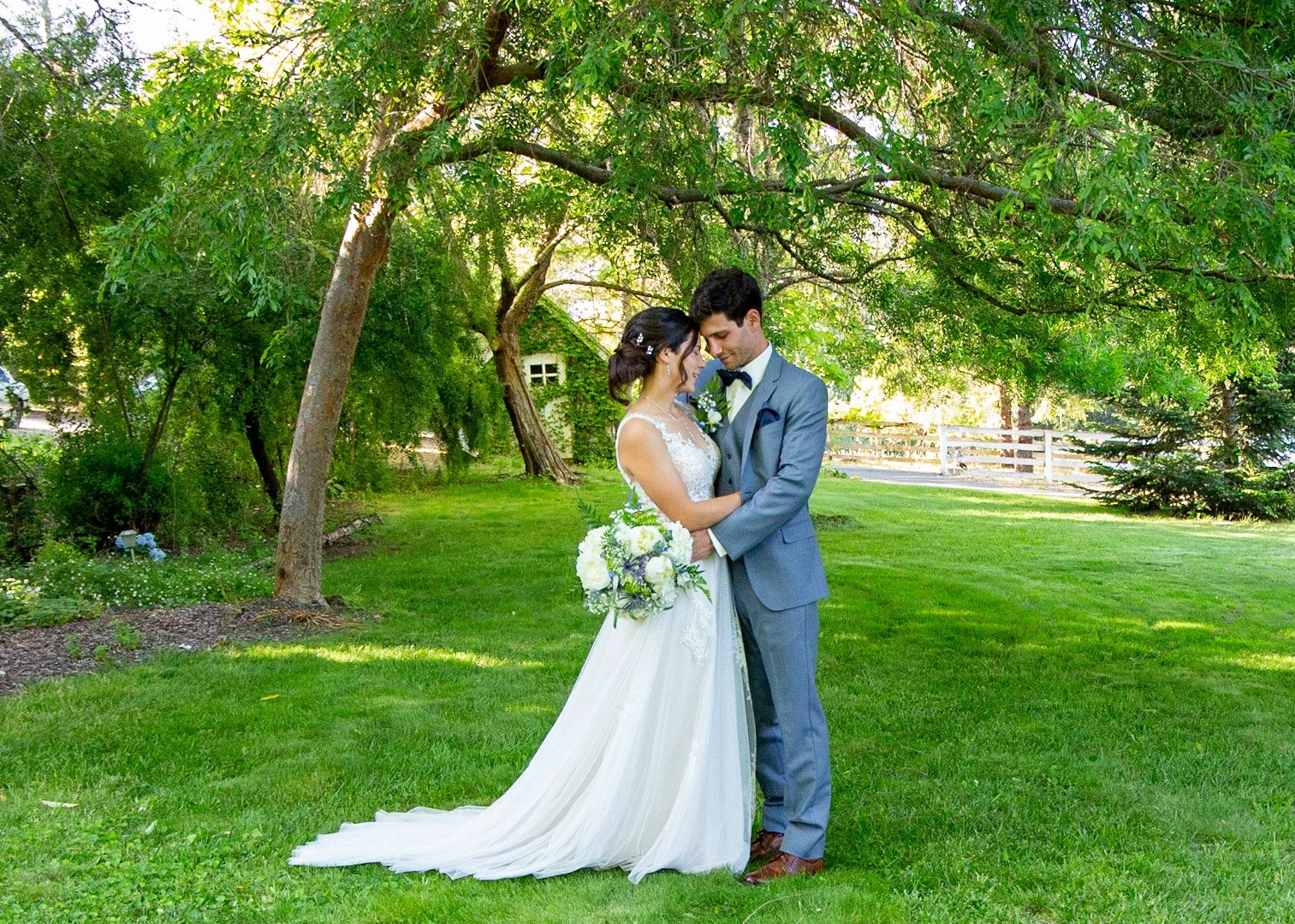 A bride and groom standing close together in a lush, green outdoor setting, sharing an intimate moment with their foreheads touching, under the shade of a large tree.