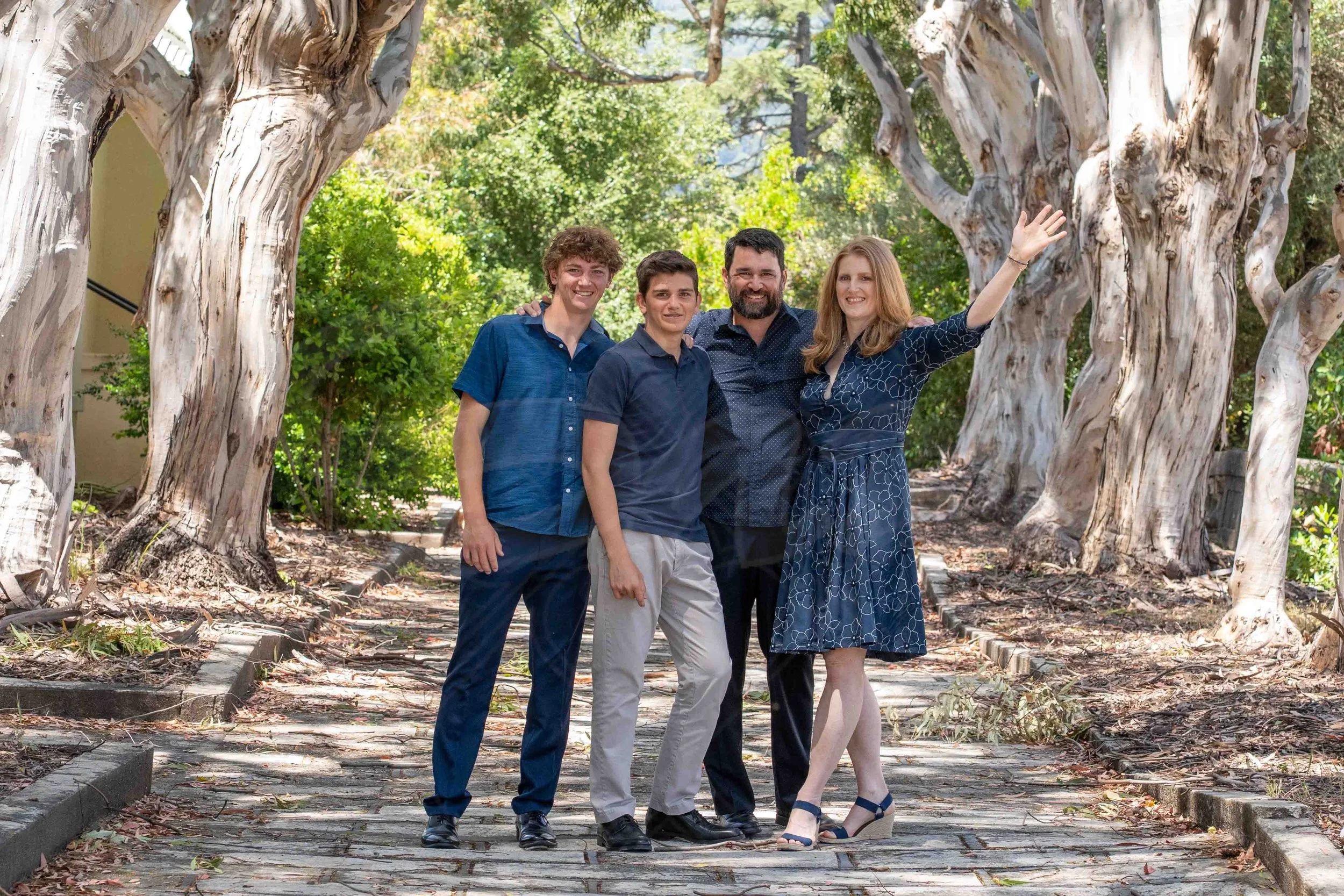A family of four standing on a cobblestone pathway in a park with large, twisted trees and green foliage, smiling and posing for the photo, with the woman waving.
